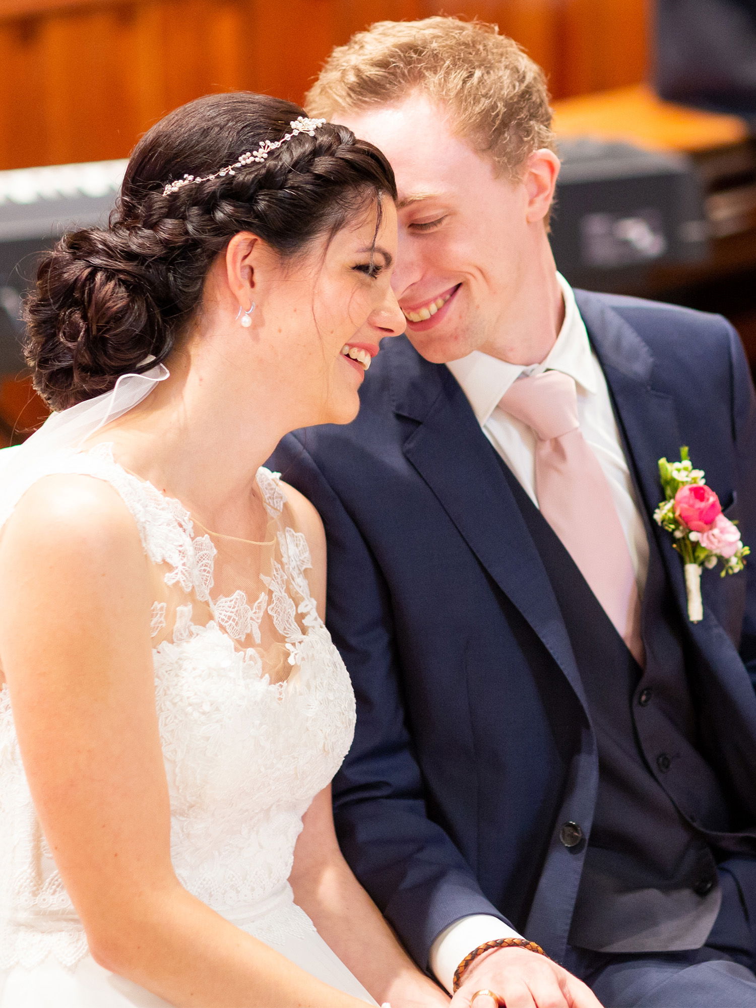 Die Braut und der Bräutigam sitzen während der Hochzeit nebeneinander und tauschen einen liebevollen Blick aus. Die Braut trägt ein weißes Kleid mit Spitzenbesatz und der Bräutigam einen dunkelblauen Anzug mit einer zarten rosa Krawatte. Beide wirken glücklich und entspannt.