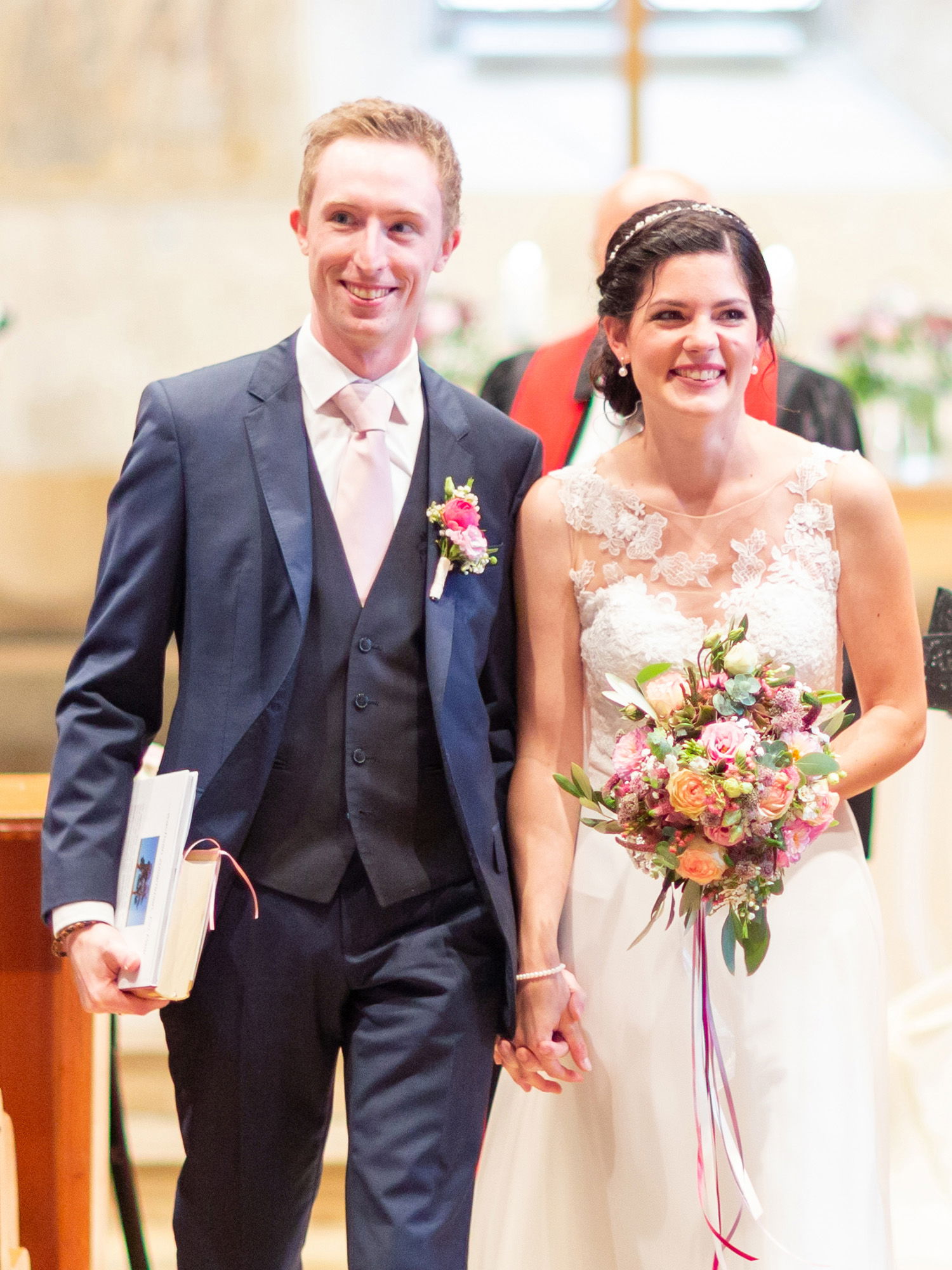 Das Brautpaar lächelt, während es nach der Zeremonie den Mittelgang hinuntergeht. Die Braut trägt ein florales Spitzenkleid und hält einen farbenfrohen Blumenstrauß. Der Bräutigam in einem eleganten Anzug trägt ein Buch in der Hand und hält die Hand seiner Braut.