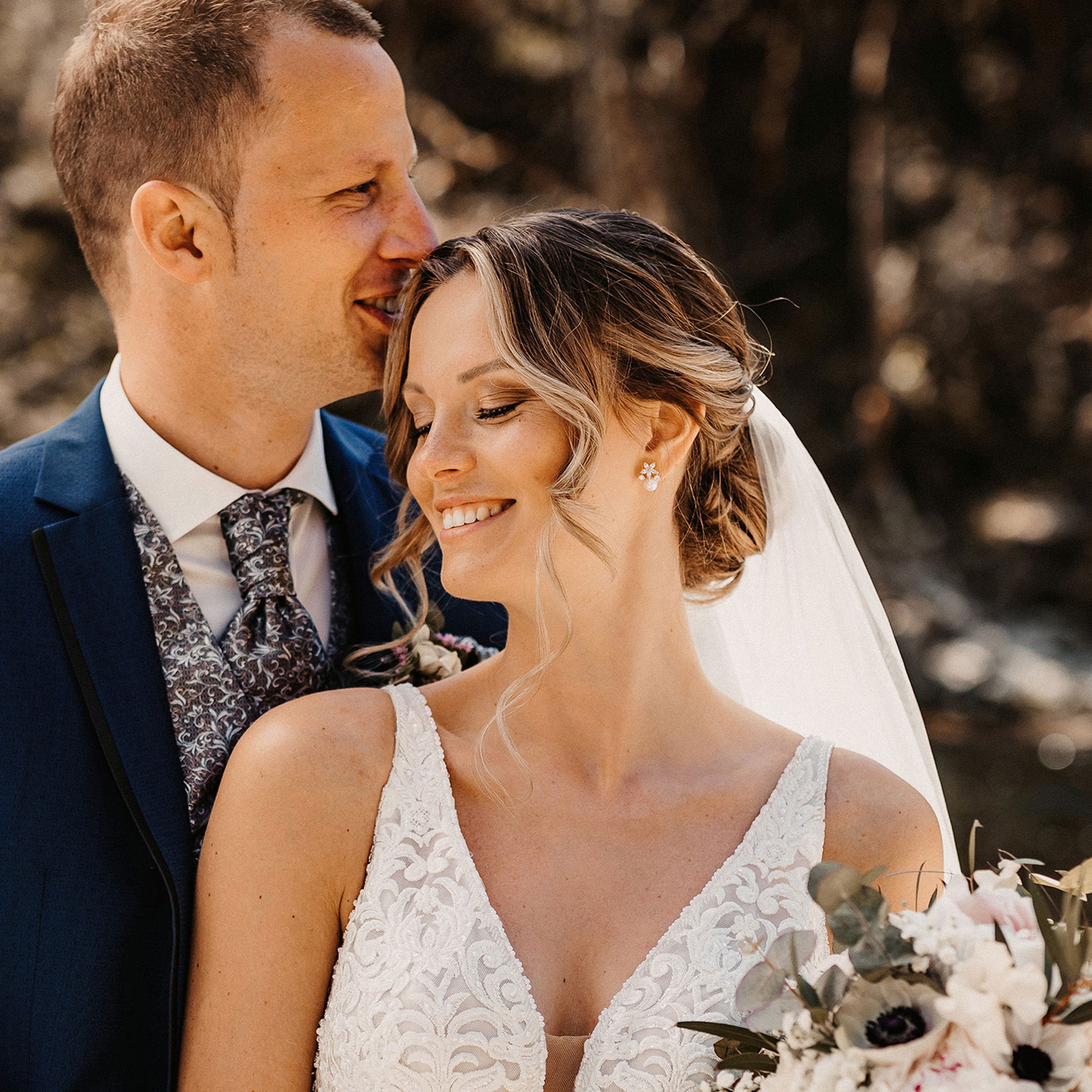 Ein nahes Portrait eines Brautpaares, der Bräutigam umarmt die Braut von hinten und flüstert ihr etwas ins Ohr, während sie zufrieden lächelt und ihren Blumenstrauß hält.