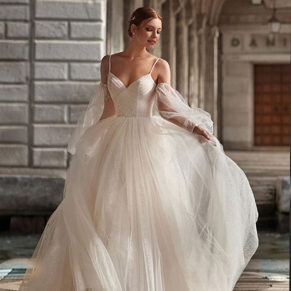 Eine Braut in einem fließenden, ärmellosen Brautkleid aus Tüll schreitet an einem historischen Gebäude entlang. Sie hält den Rock ihres Kleides und blickt sanft nach unten, während der Wind durch den Stoff weht.