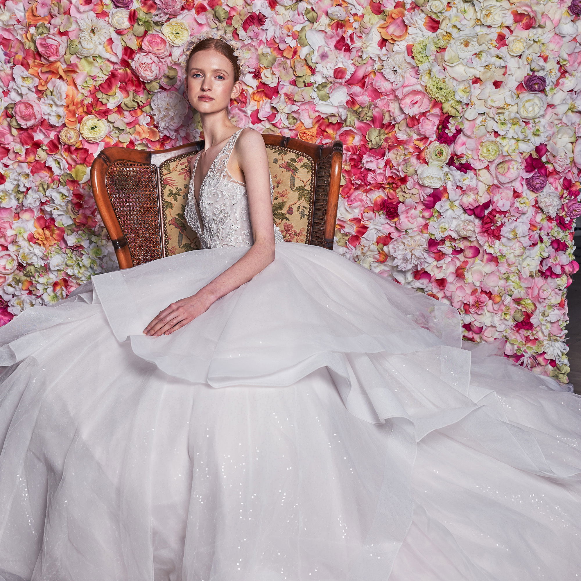 Eine Braut mit einem weit ausladenden, mehrlagigen Brautkleid sitzt auf einem antiken Stuhl vor einer blühenden Wand aus rosa und cremefarbenen Ranunkeln. Das Kleid wirkt majestätisch, und die Blumen im Hintergrund verstärken den romantischen Charme der Szene.
