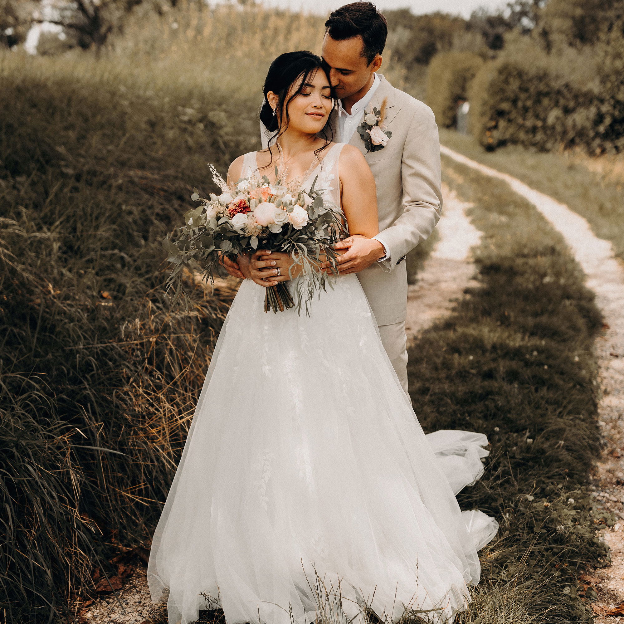 Die Braut, in einem weißen, eleganten Kleid, hält einen zarten Blumenstrauß, während der Bräutigam in einem beigen Anzug liebevoll hinter ihr steht. Sie befinden sich auf einem Pfad, umgeben von Natur.