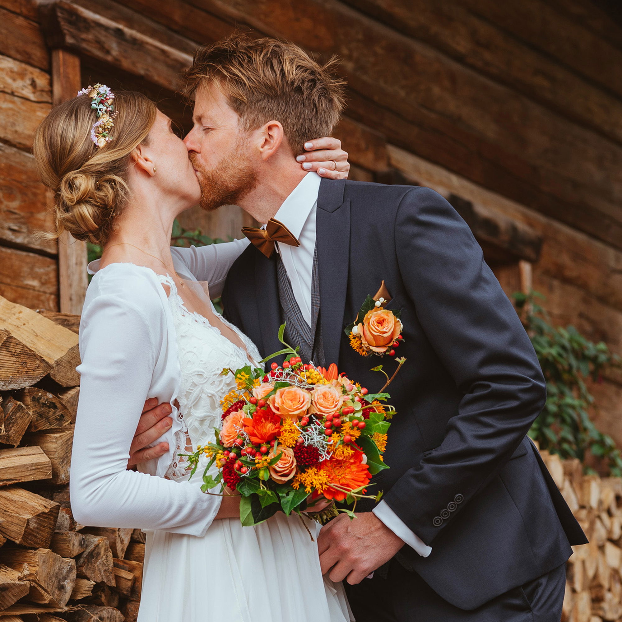 Ein frisch verheiratetes Paar küsst sich leidenschaftlich vor einem Stapel Holzscheite. Die Braut hält einen farbenfrohen Blumenstrauß in warmen Orangetönen, und der Bräutigam trägt einen eleganten, dunklen Anzug mit einer braunen Fliege.
