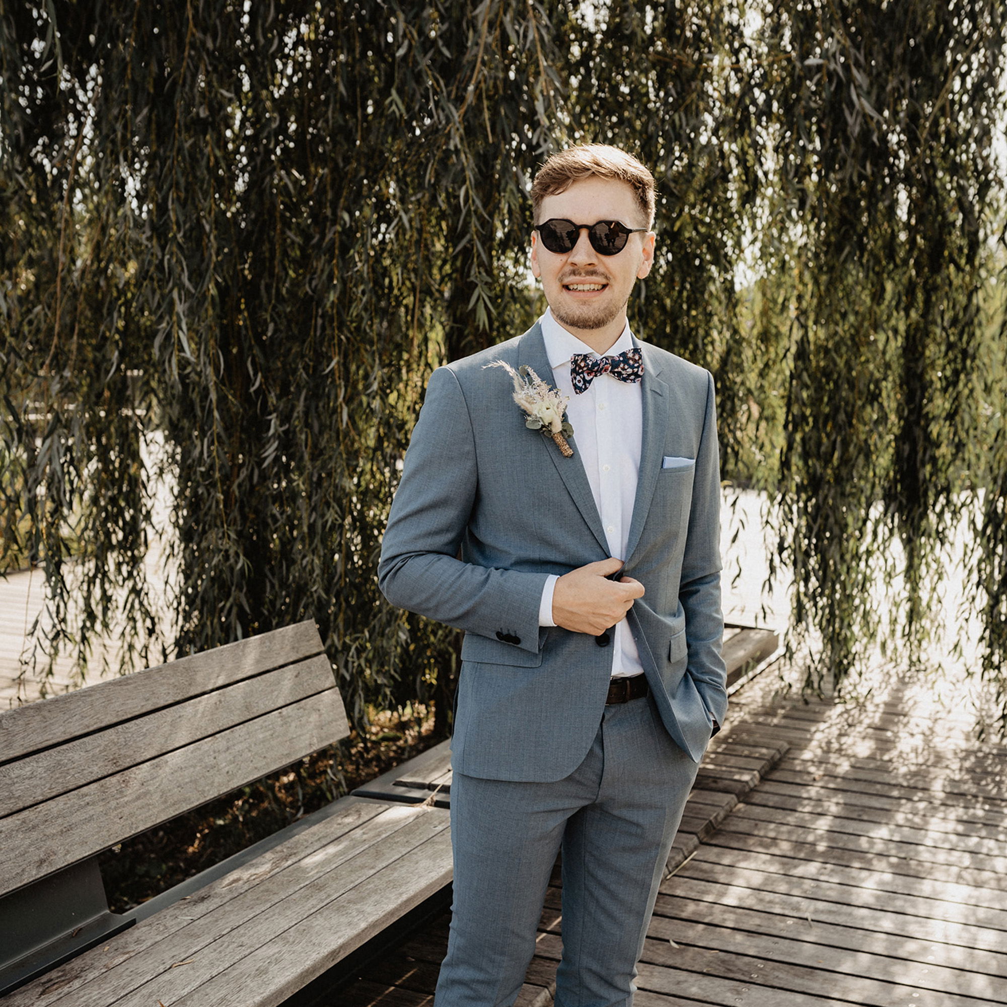 Ein Mann in einem eleganten grauen Anzug und modischer Sonnenbrille steht lässig auf einem Holzsteg, umgeben von herabhängenden Weidenblättern. Er trägt eine gemusterte Fliege und lächelt selbstbewusst in die Kamera.
