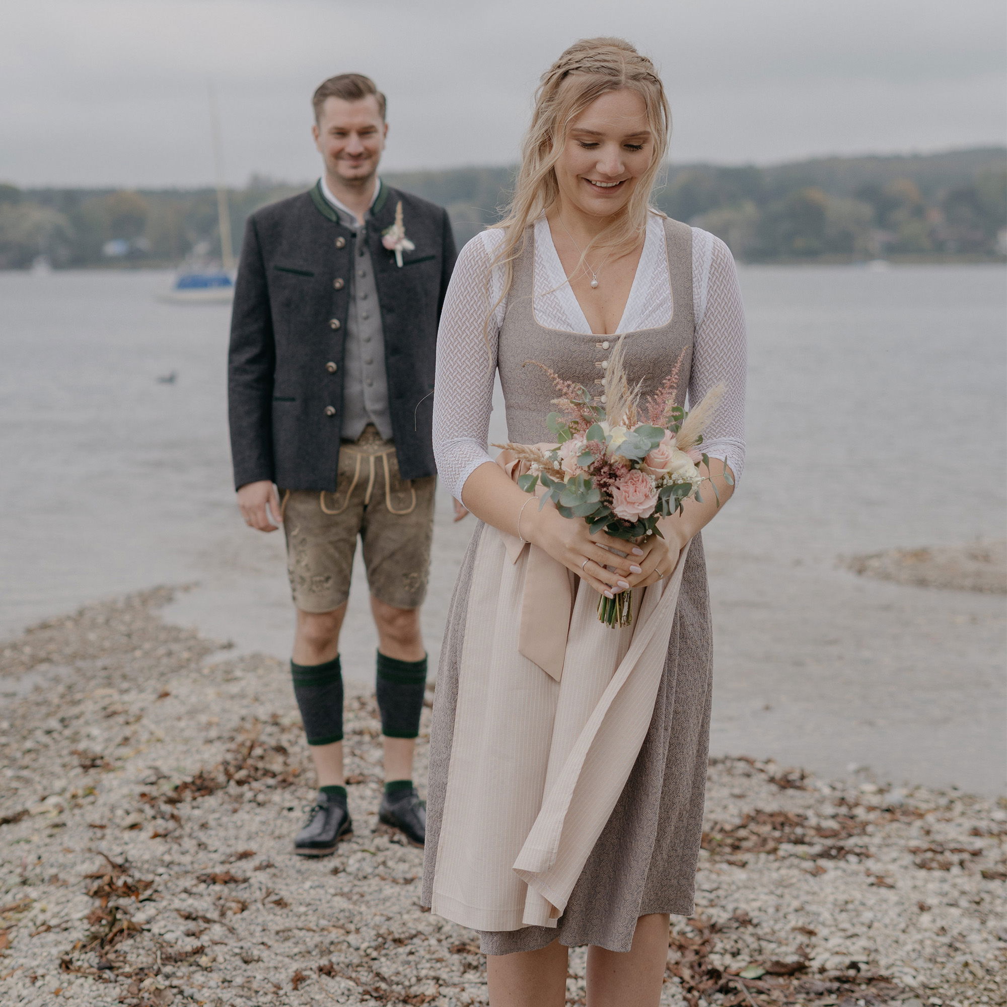 Eine Braut in einem eleganten Dirndl blickt lächelnd auf ihren Blumenstrauß, während sie am Seeufer steht. Der Bräutigam in Lederhosen und Tracht nähert sich lächelnd von hinten.