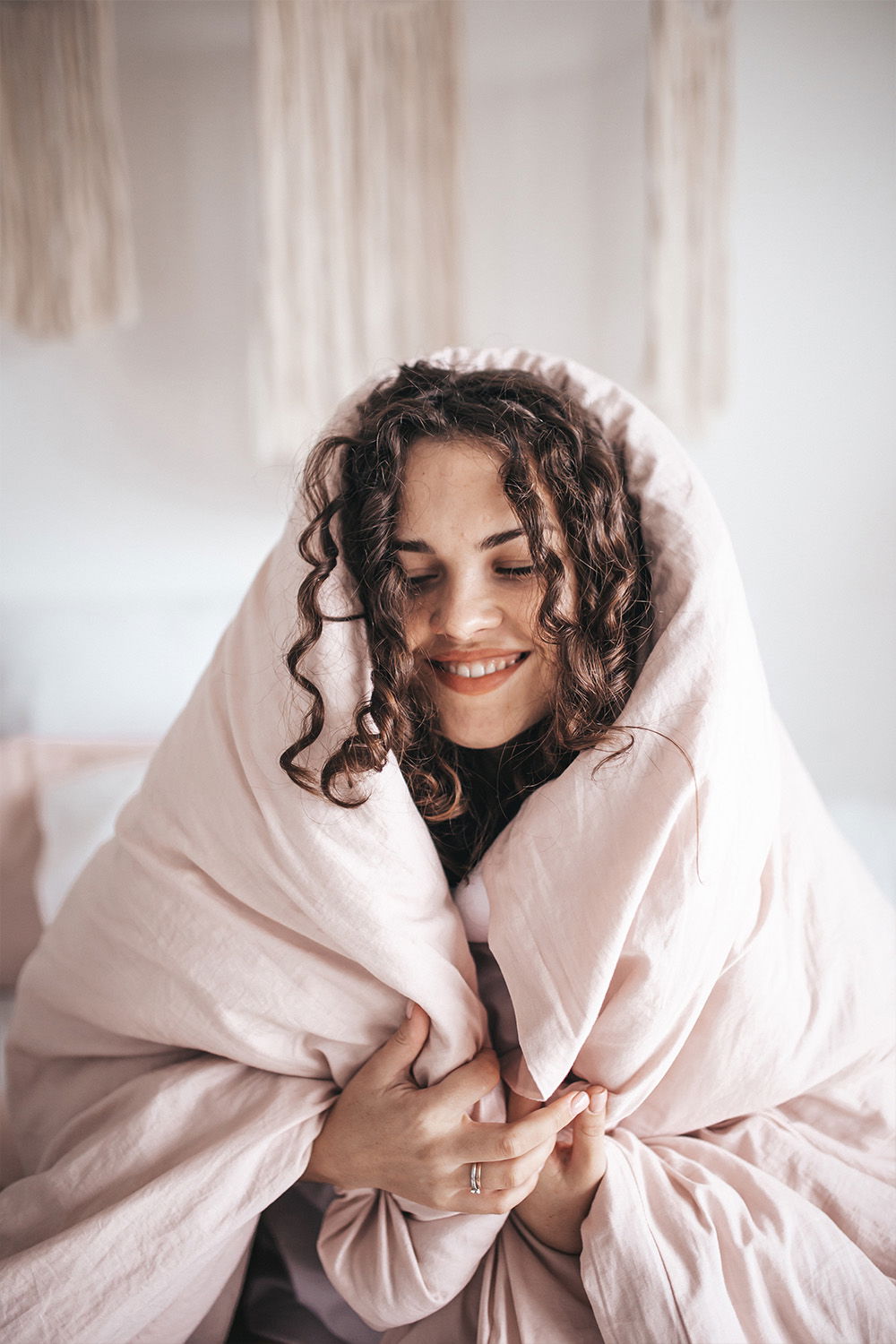 Eine Frau sitzt lächelnd in einer weichen, beigen Decke eingewickelt. Ihr lockiges Haar umrahmt ihr Gesicht, und sie wirkt entspannt und zufrieden. Das Bild vermittelt ein Gefühl von Geborgenheit und Entspannung.