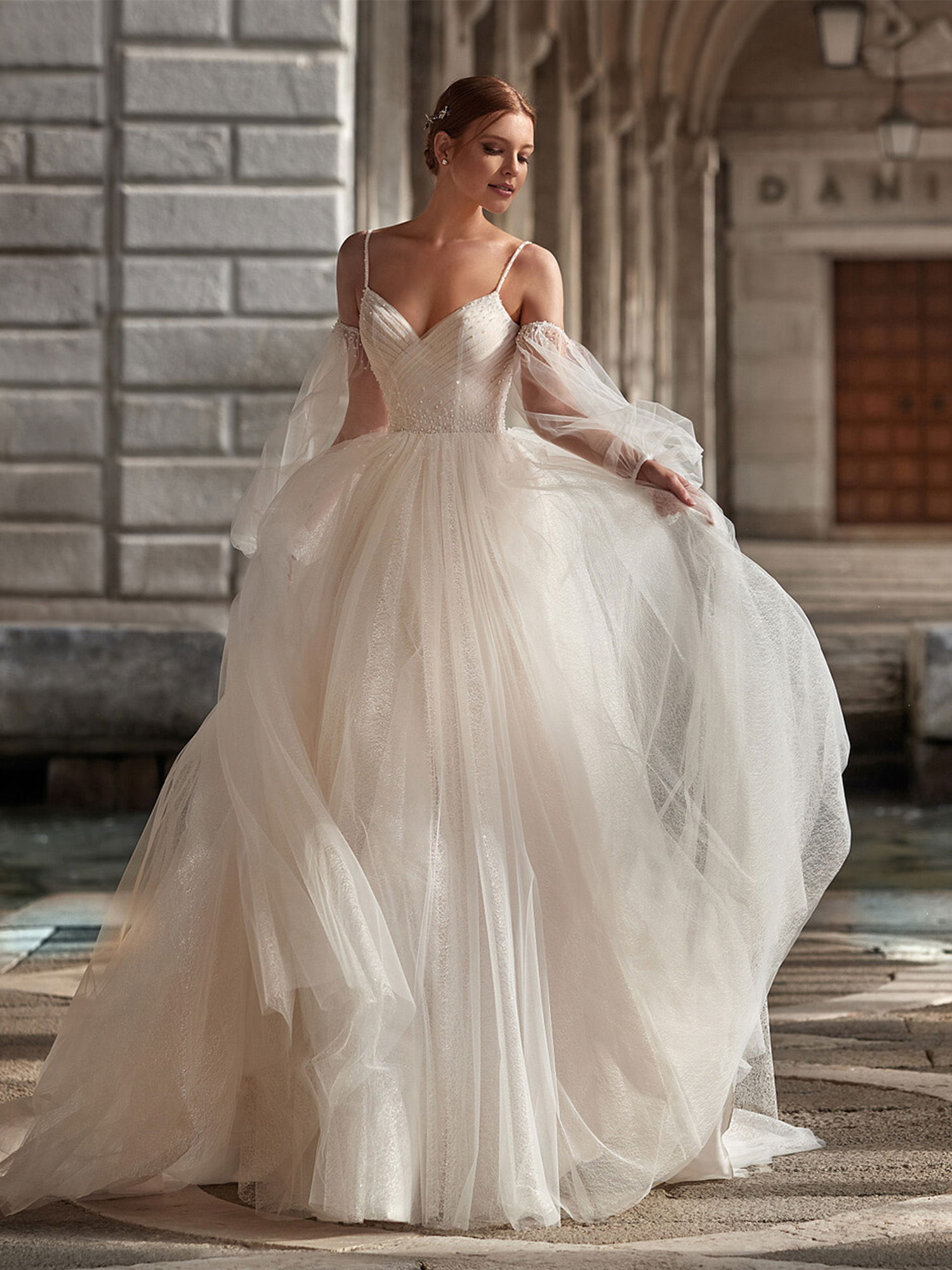 Eine Frau in einem fließenden Brautkleid aus Tüll posiert an einem alten Steinbau. Ihr Kleid hat schmale Träger und abnehmbare Tüllärmel, die dem Look eine verträumte Note verleihen. Sie schaut sanft nach unten und genießt den Moment.