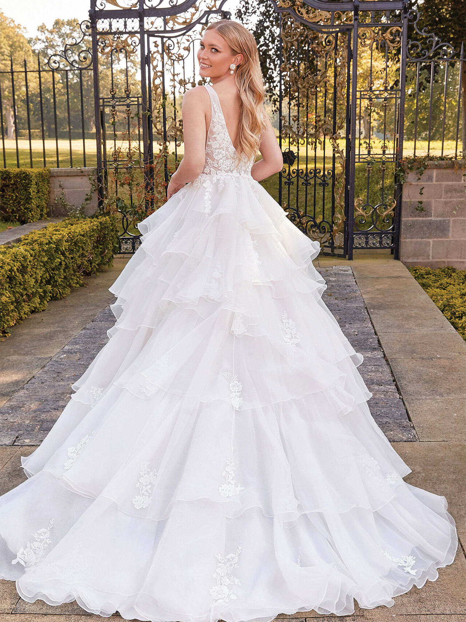 Die Braut trägt ein voluminöses Duchesse-Brautkleid mit floral besticktem Mieder und mehreren Tülllagen. Sie steht vor einem Tor in einem gepflegten Garten und dreht sich leicht, während sie über die Schulter lächelt.