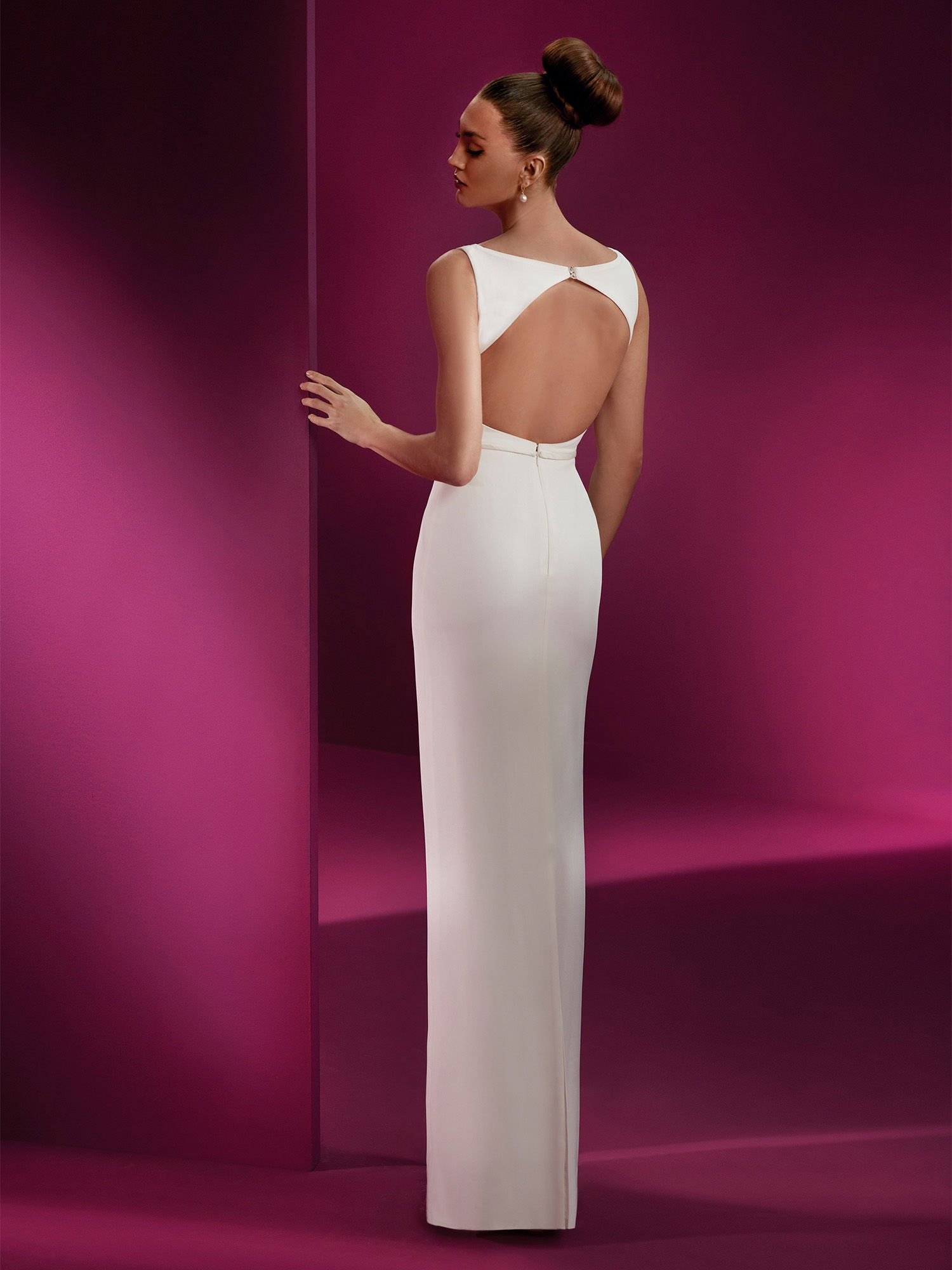 Ein Model trägt ein weißes, figurbetontes Kleid mit einem dramatischen Rückenausschnitt. Sie steht vor einem tiefvioletten Hintergrund, und das Design des Kleides betont ihre schlanke Silhouette und den minimalistischen Stil.