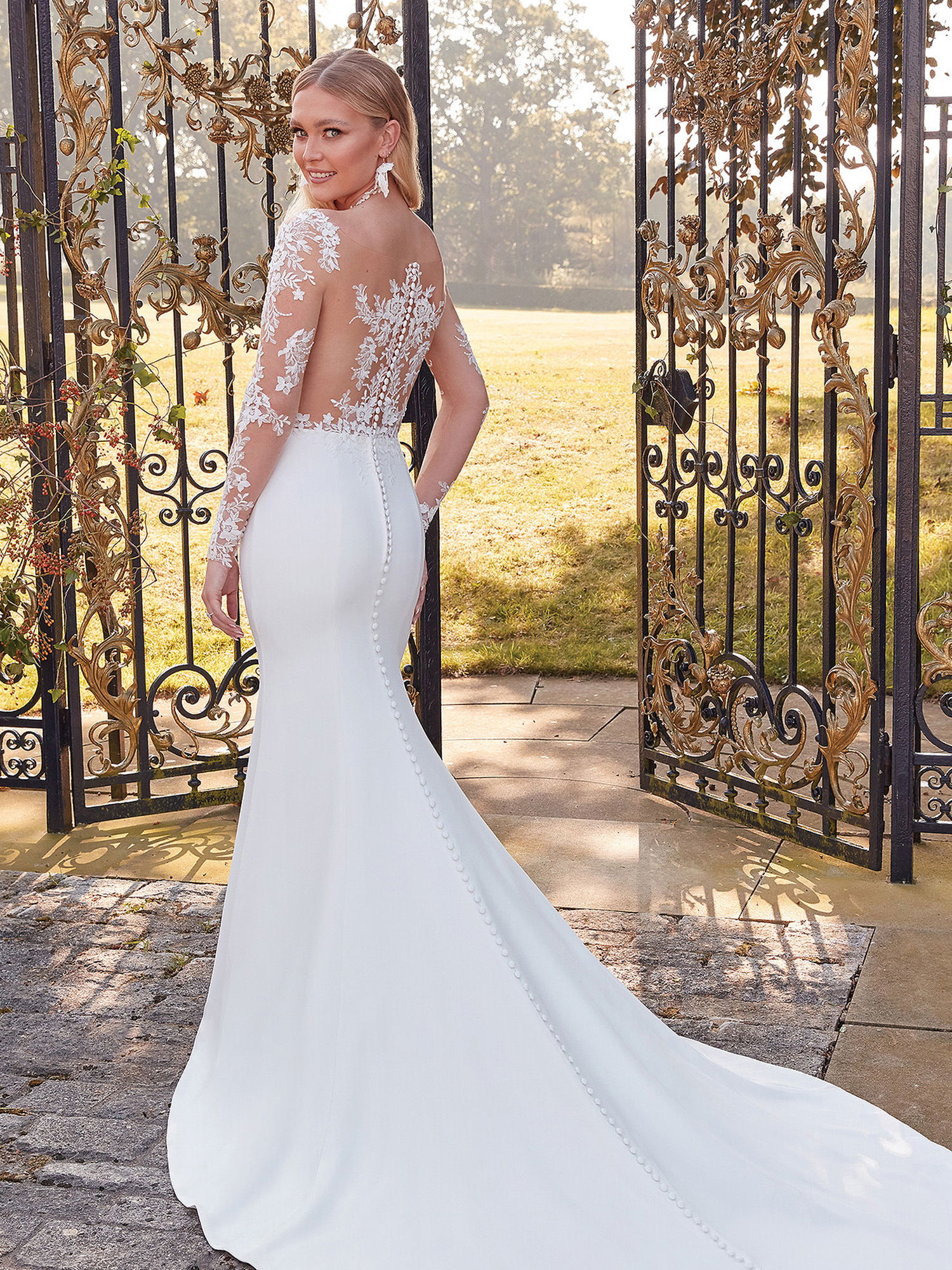 Eine Frau trägt ein figurbetontes Godet-Hochzeitskleid mit floralen Spitzenärmeln und einem transparenten Rückenausschnitt. Sie posiert vor einem kunstvollen Eisentor und lächelt, während der Stoff ihres Kleides sanft hinter ihr fällt.