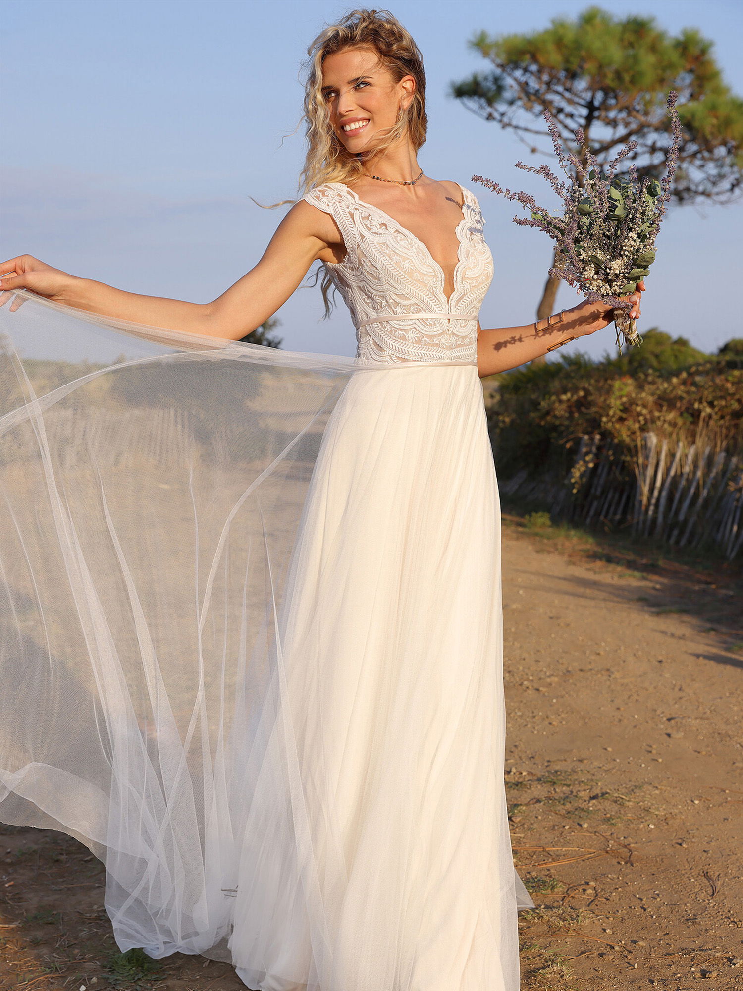 Eine Frau in einem romantischen, ärmellosen Hochzeitskleid mit Spitzenverzierung und einem leichten Tüllrock steht in einer naturnahen Umgebung. Sie hält einen Blumenstrauß und lächelt, während der Wind sanft durch den Stoff weht.
