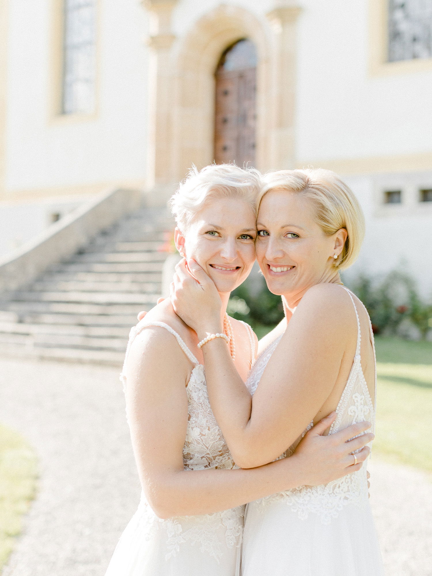 Zwei glückliche Bräute stehen in eleganten, weißen Hochzeitskleidern vor einem antiken Gebäude. Ihre Umarmung und ihr herzliches Lächeln vermitteln eine tiefe Verbundenheit, während sie diesen besonderen Moment im strahlenden Sonnenlicht festhalten.