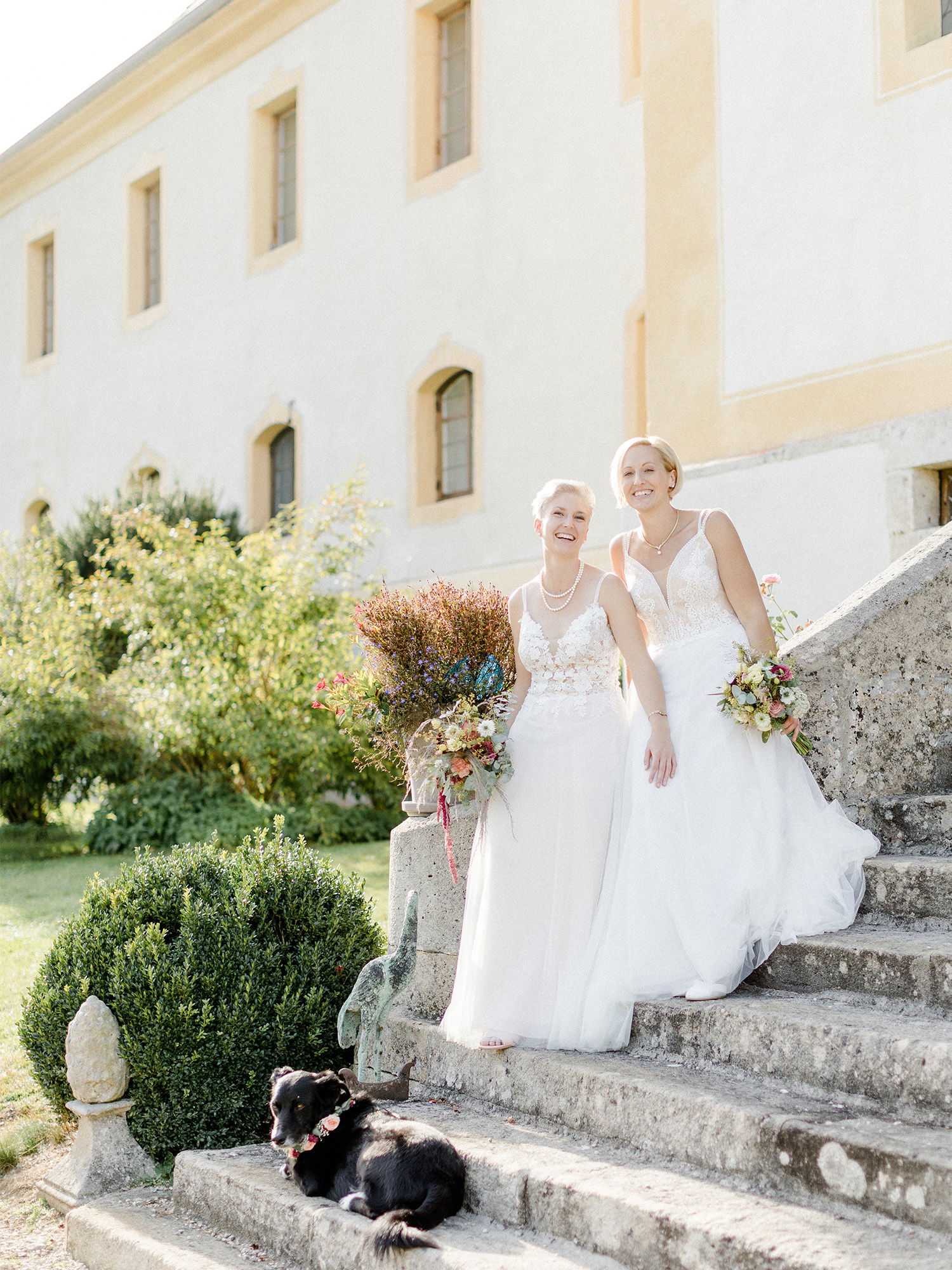 Die Bräute in eleganten, weißen Hochzeitskleidern sitzen entspannt auf den Stufen eines alten Gebäudes. Beide halten farbenfrohe Blumensträuße, während ein schwarzer Hund neben ihnen auf der Treppe ruht und den Moment mit ihnen teilt.