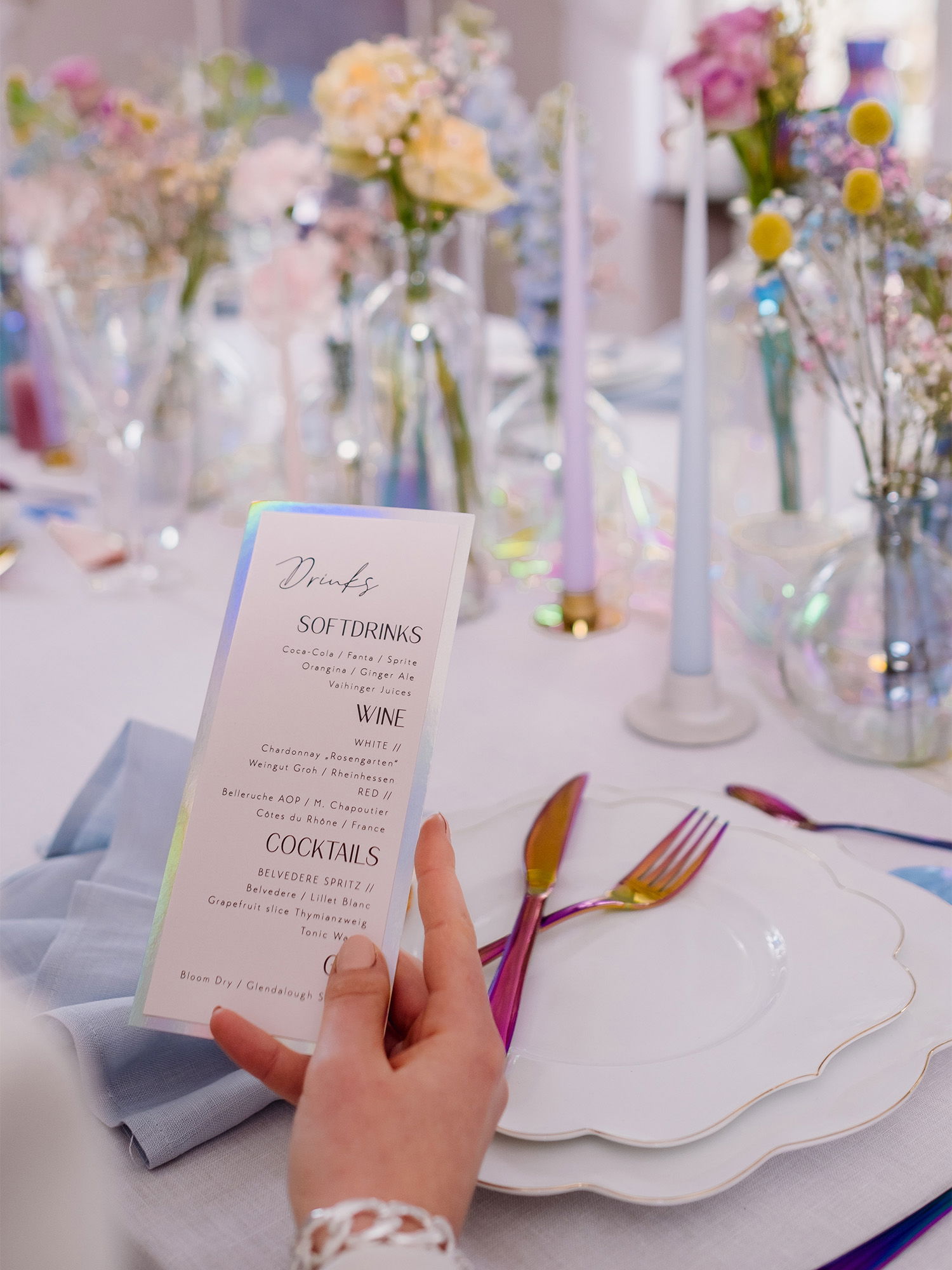 Eine Nahaufnahme einer Hand, die eine Getränkekarte auf einem stilvollen Tisch präsentiert. Der Tisch ist mit Glasvasen voller Blumen in sanften Farben und dezenten, modernen Kerzen geschmückt. Das Besteck glänzt metallisch bunt, passend zum feierlichen Ambiente.