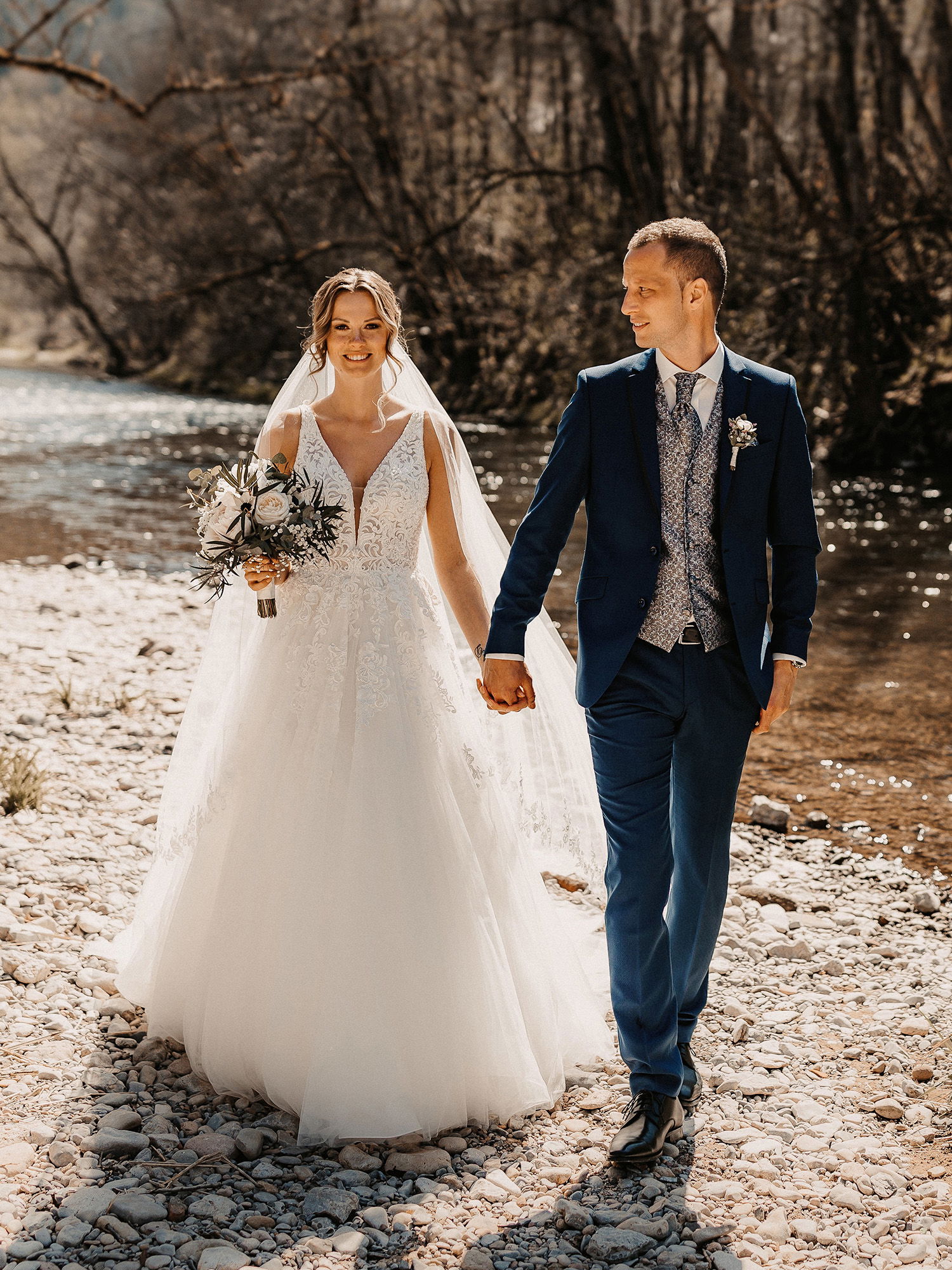 Das frisch verheiratete Paar geht über einen Kiesweg am Flussufer, die Braut strahlt in ihrem weißen Hochzeitskleid, während der Bräutigam sie lächelnd anschaut.