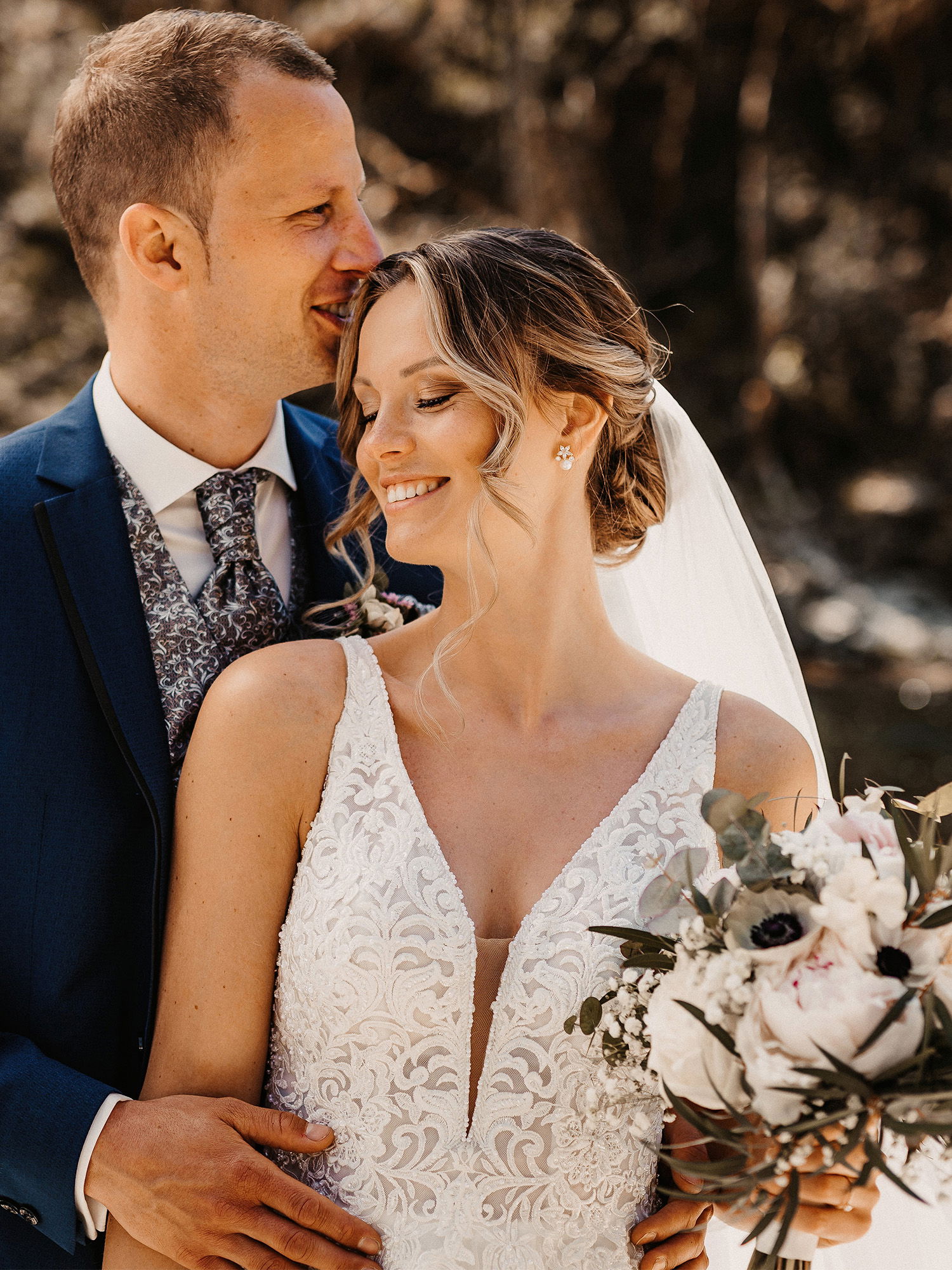 Das Brautpaar steht eng umschlungen, der Bräutigam lehnt seinen Kopf sanft an den der Braut, die ein strahlendes Lächeln und einen stilvollen Blumenstrauß zeigt.