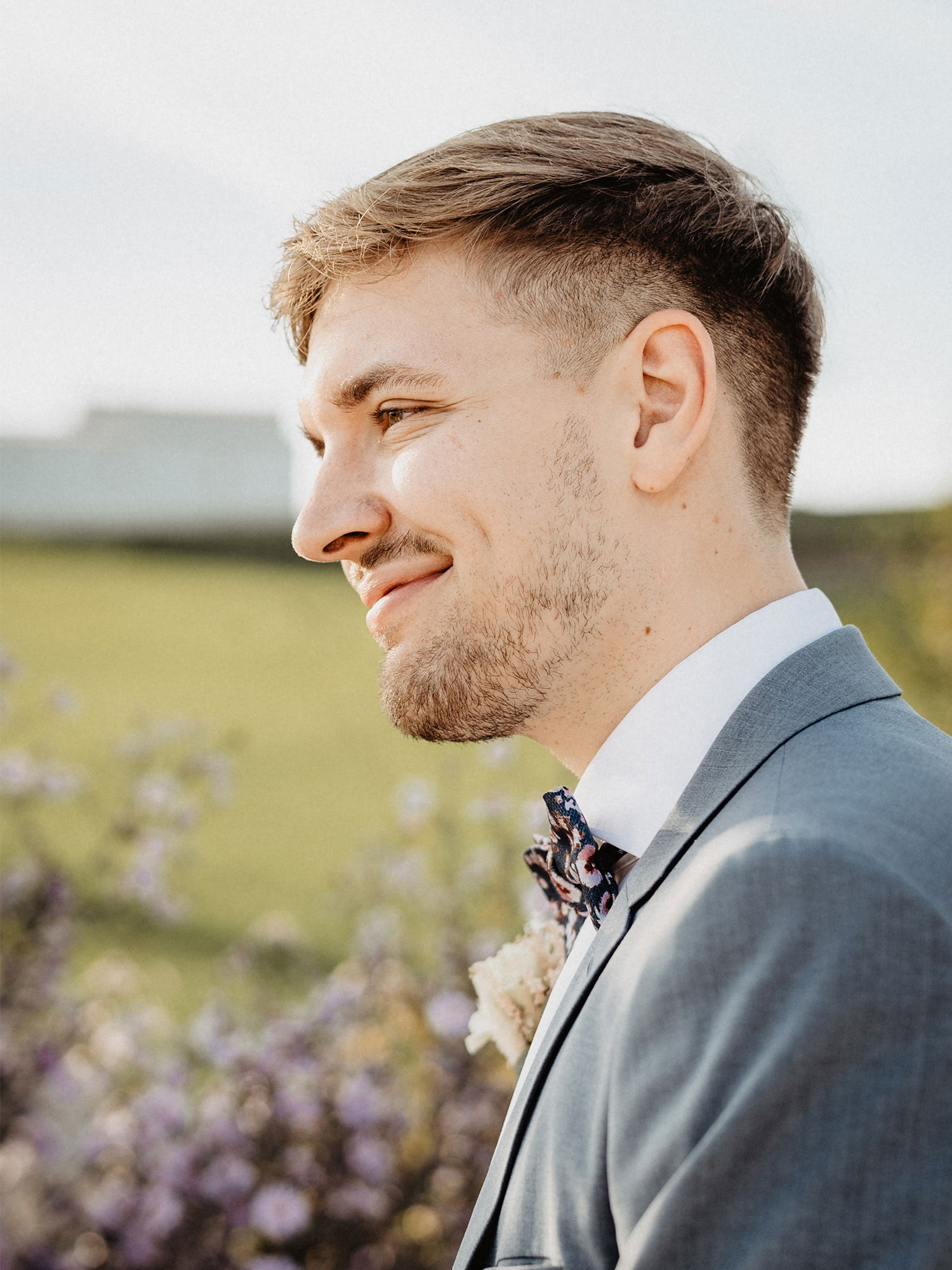 Nahaufnahme eines lächelnden Mannes im grauen Anzug mit einer auffälligen Fliege. Der Hintergrund zeigt eine ländliche Landschaft mit Blumen im sanften Licht.