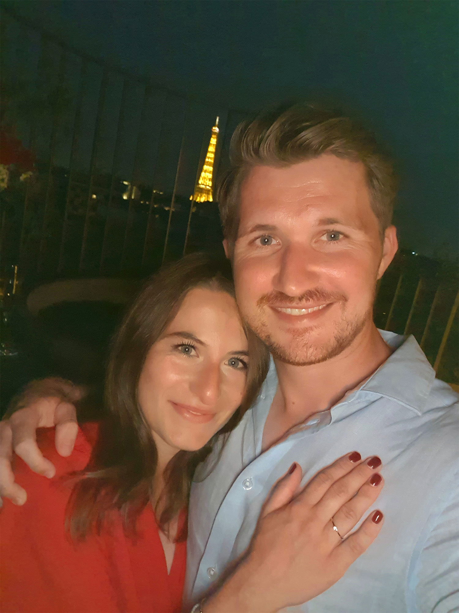 Ein frisch verlobtes Paar lächelt in die Kamera vor einer nächtlichen Kulisse in Paris. Der Eiffelturm ist im Hintergrund zu sehen, und die Frau zeigt glücklich ihren Verlobungsring.