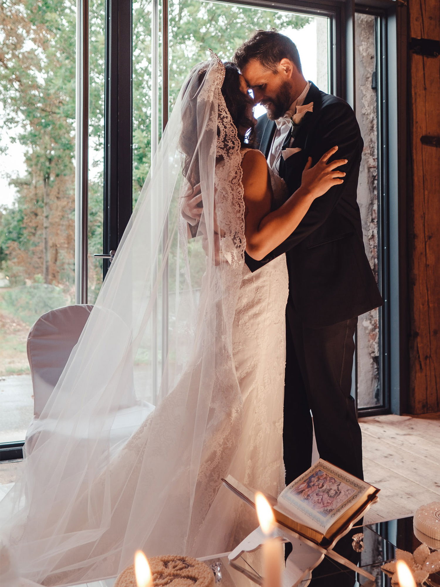 Die Braut und der Bräutigam stehen eng umschlungen vor einem großen Fenster, das den Blick auf die Natur freigibt. Die Braut trägt ein romantisches Kleid mit Schleier und der Bräutigam einen eleganten Anzug. Im Vordergrund sind Kerzen und ein religiöses Buch platziert.