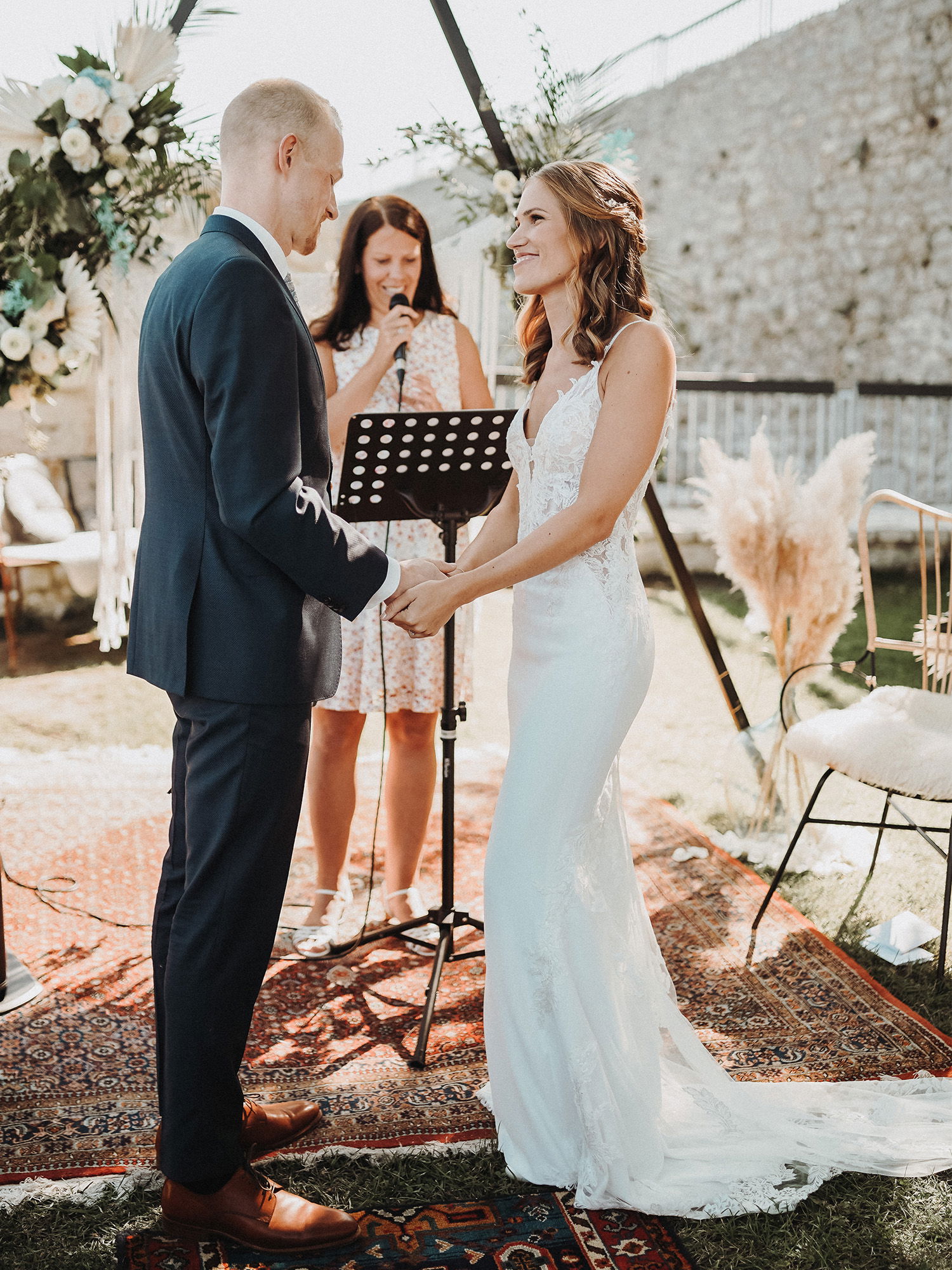 Die Braut und der Bräutigam stehen während ihrer Hochzeit auf einem dekorativen Teppich und halten sich die Hände. Die Braut trägt ein enges Hochzeitskleid mit Spitze und der Bräutigam ist in einem eleganten, dunklen Anzug gekleidet. Eine Traurednerin spricht in ein Mikrofon hinter ihnen.