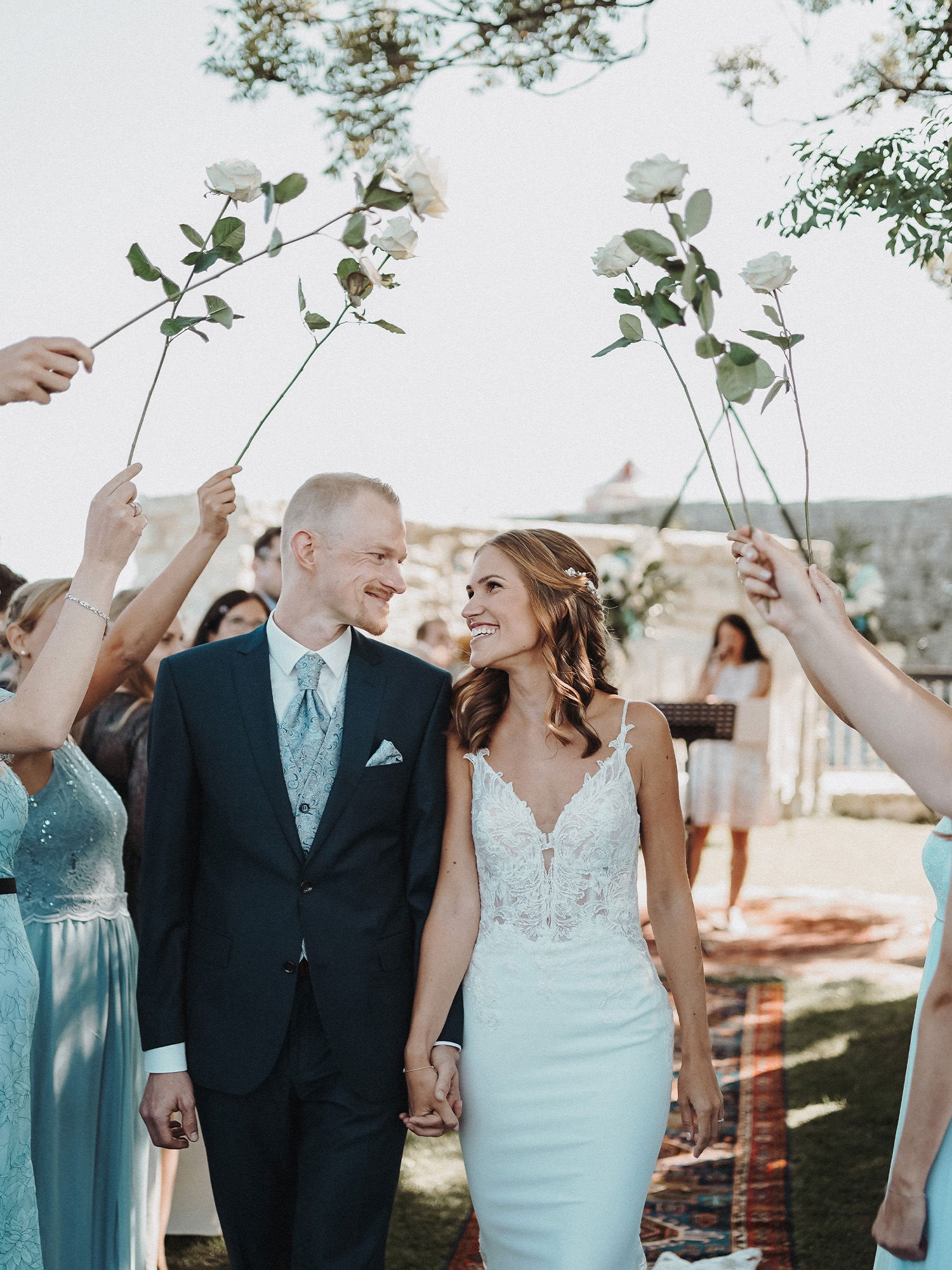 Nach der Trauung geht das frisch verheiratete Paar Hand in Hand durch eine Gasse aus erhobenen Rosen, die von den Hochzeitsgästen gehalten werden. Die Braut strahlt in einem figurbetonten Spitzenkleid, während der Bräutigam in einem schicken Anzug an ihrer Seite geht.
