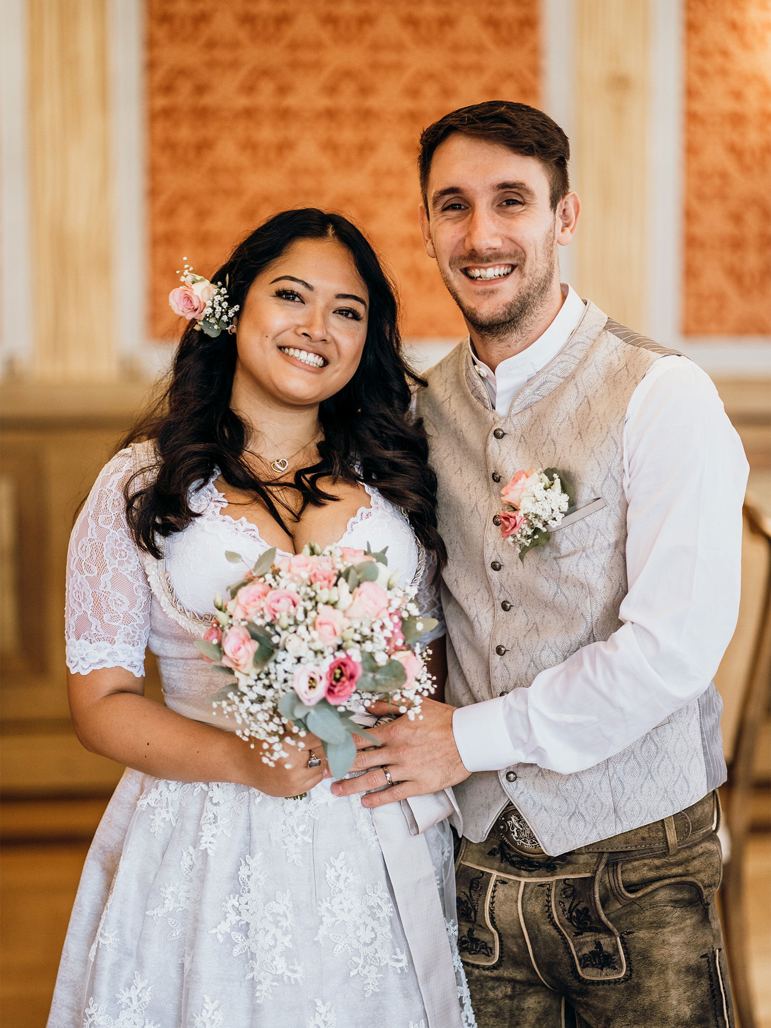 Ein frisch verheiratetes Paar posiert für ein Foto in einem festlich dekorierten Raum. Die Braut hat ein elegantes Dirndl mit Spitze an und der Bräutigam trägt eine Tracht mit Weste und Lederhosen. Sie strahlen vor Freude.