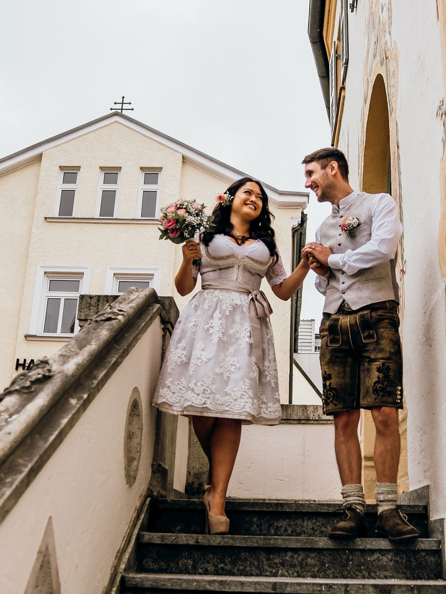Ein Brautpaar schreitet lächelnd eine Treppe hinunter. Die Braut trägt ein schickes, weißes Kleid mit floralen Mustern, und der Bräutigam ist in eine traditionelle Lederhose gekleidet. Beide wirken glücklich und verbunden, während sie Hand in Hand gehen.