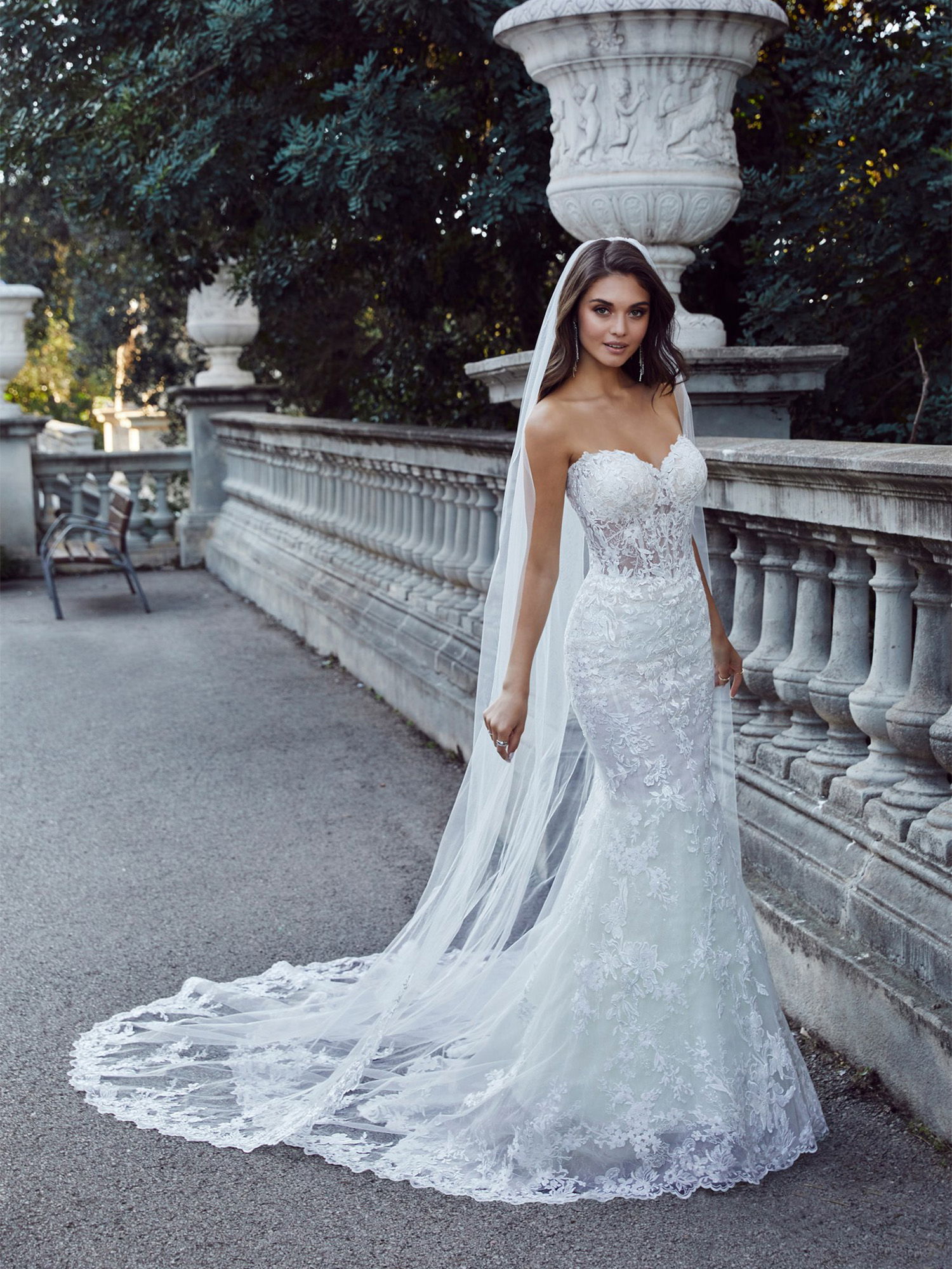 Eine Frau in einem schulterfreien Mermaid-Kleid mit aufwendiger Spitze und einem langen, bestickten Schleier posiert an einer steinernen Balustrade. Der enge Schnitt des Kleides betont ihre Figur und verleiht dem Look eine glamouröse Note.
