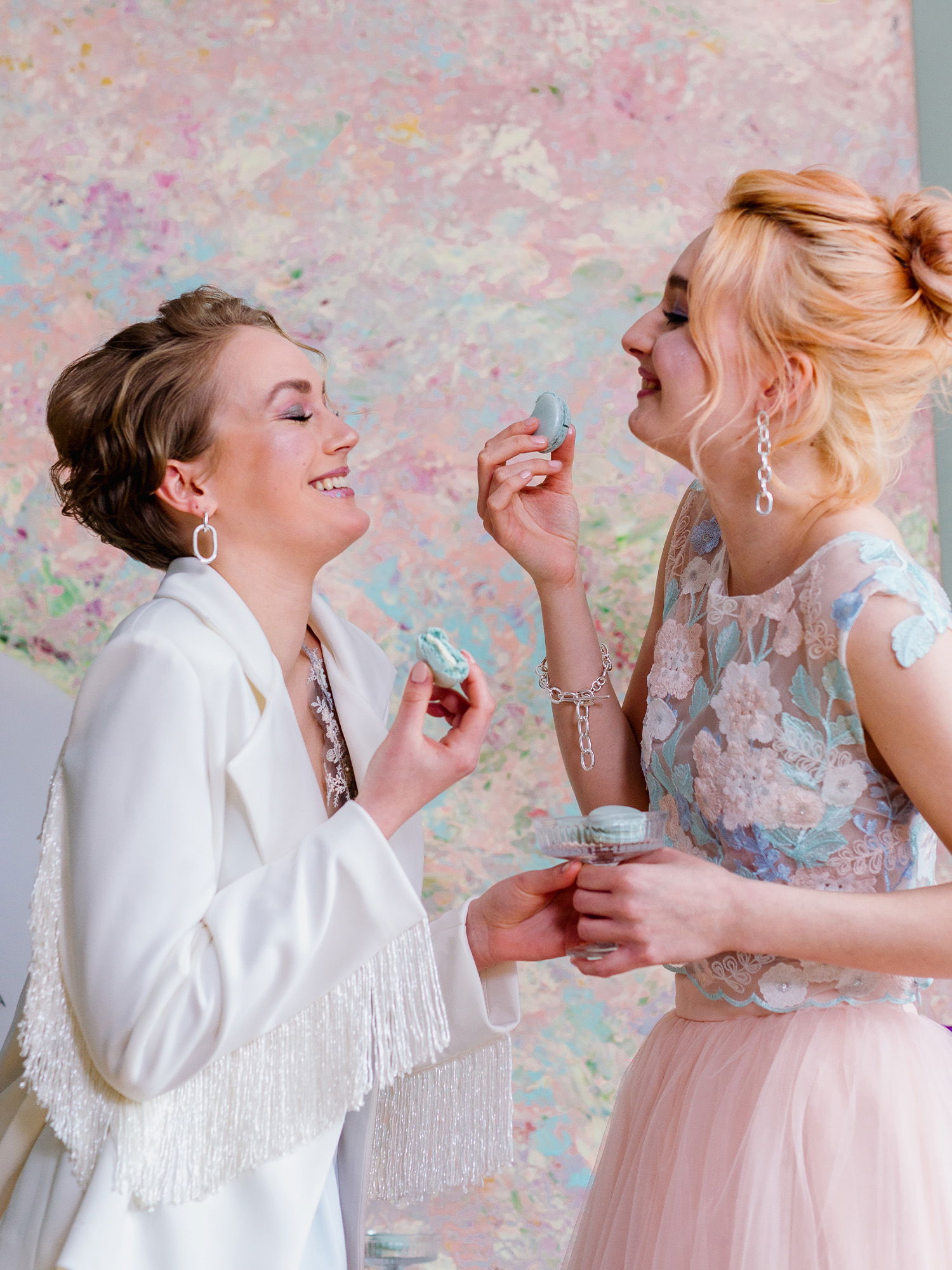 Zwei Freundinnen genießen gemeinsam Macarons und lachen herzlich. Eine trägt einen weißen Blazer mit Fransen und die andere ein helles Spitzenkleid. Der pastellfarbene Hintergrund mit abstrakten Mustern verleiht der Szene eine verspielte, künstlerische Atmosphäre.