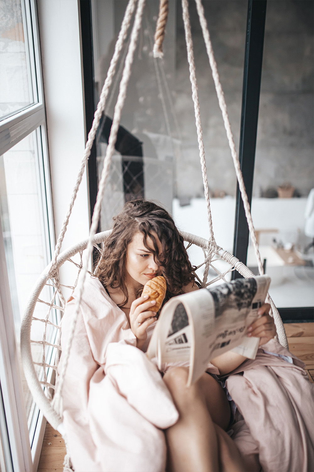 Eine Frau in einer Hängeschaukel mit einer Decke liest entspannt eine Zeitung und isst ein Croissant. Sie sitzt in der Nähe eines Fensters, durch das sanftes Licht in den Raum fällt, was eine gemütliche Atmosphäre schafft.