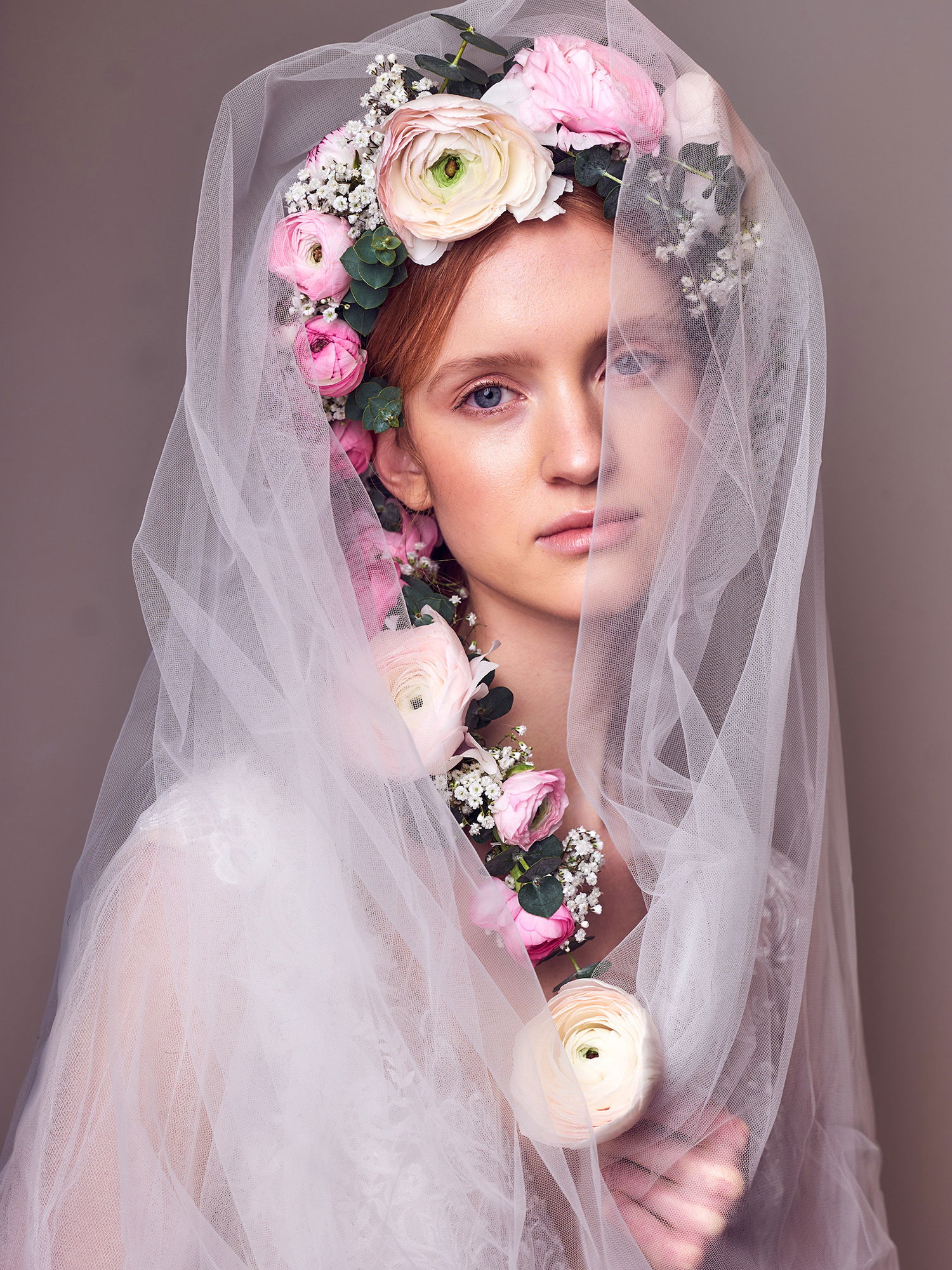 Eine Braut, deren Gesicht teilweise von einem weißen Schleier verdeckt wird, trägt einen Blütenkranz aus Ranunkeln in Rosa- und Cremetönen. Der florale Kopfschmuck, kombiniert mit dem zarten Schleier, erzeugt ein verträumtes, romantisches Bild.