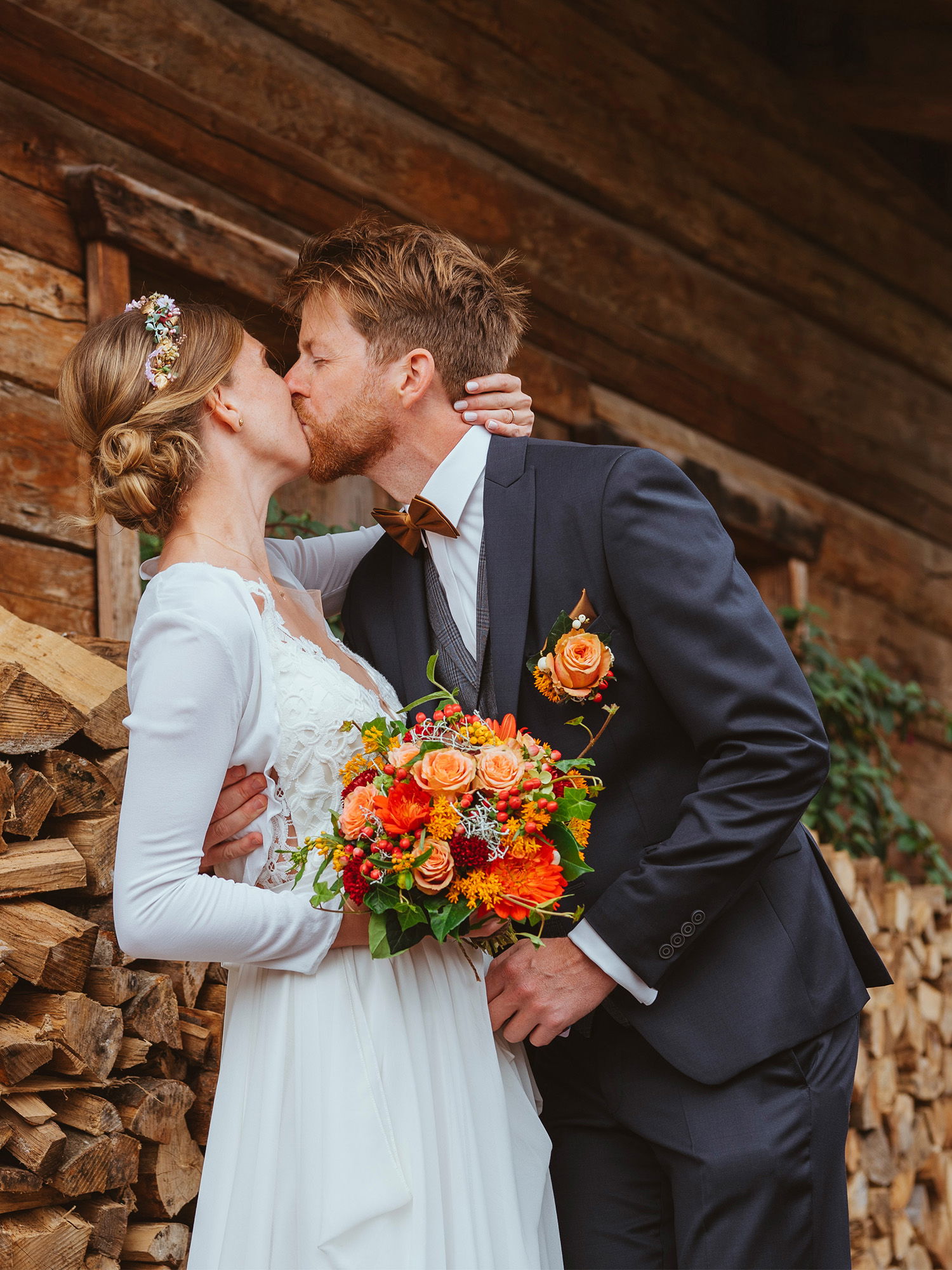 Eine Braut und ein Bräutigam küssen sich vor einem Stapel Holzscheite. Die Braut trägt ein langes, elegantes Kleid und einen bunten Blumenstrauß, der Bräutigam ist in einem stilvollen Anzug mit Fliege gekleidet.