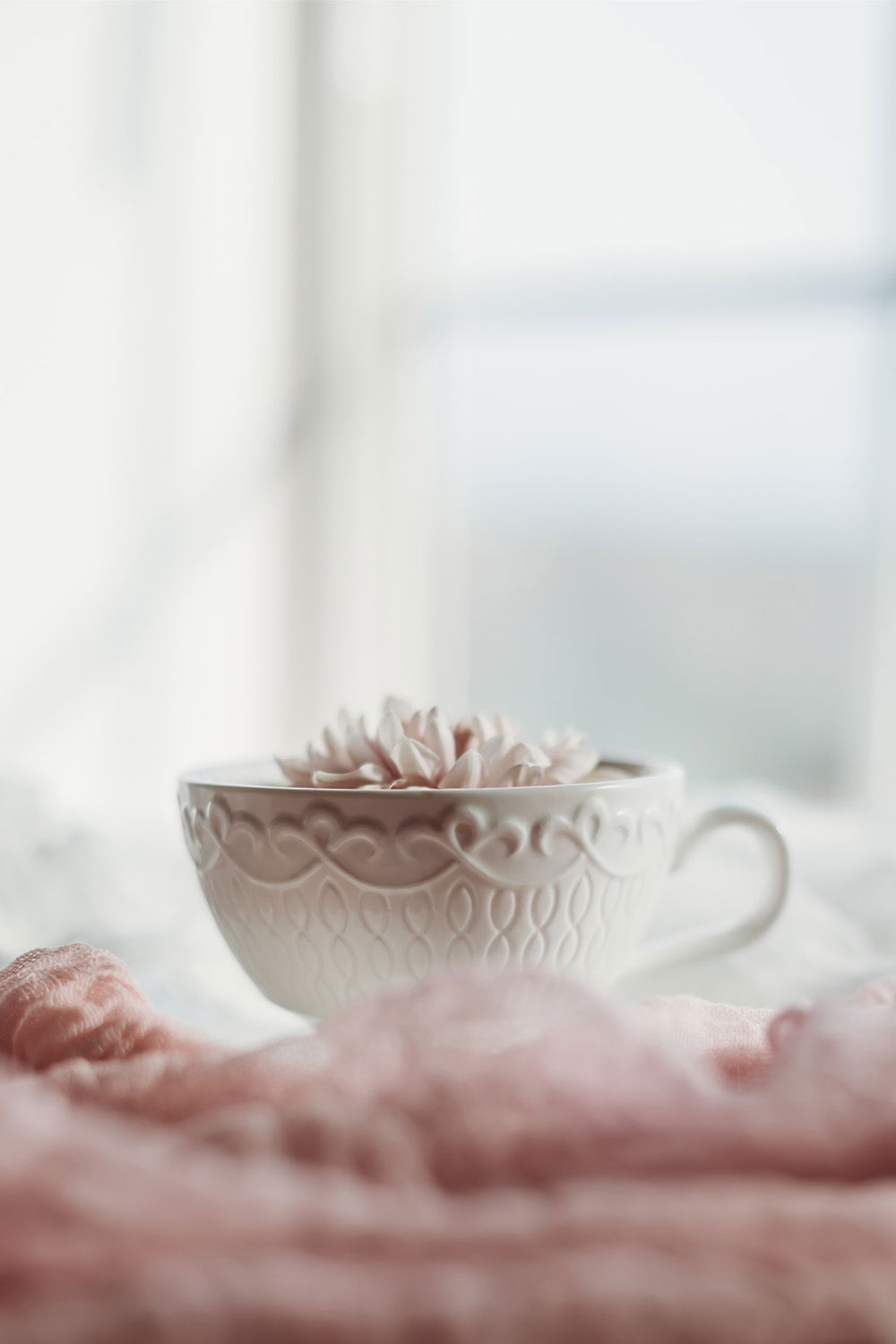Eine kunstvoll verzierte Teetasse mit einer Blume darin steht auf einem weichen, rosa Stoff. Die Szene im sanften Licht vermittelt Ruhe und Gemütlichkeit, perfekt für einen entspannten Moment.