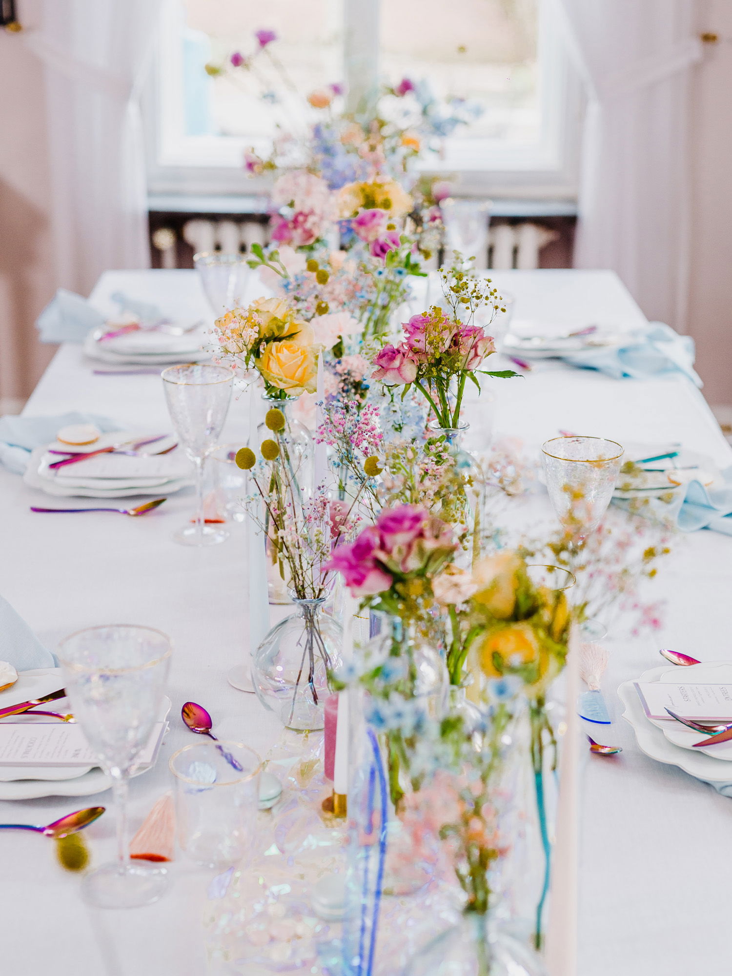 Ein festlich gedeckter Tisch mit einer langen Reihe von Glasvasen, die mit bunten Blumen in sanften Farben gefüllt sind. Durch das Fenster strömt Tageslicht und verleiht der romantischen Hochzeitsdekoration einen sanften Glanz. Das Regenbogenbesteck auf den Tellern fügt eine moderne Note hinzu.