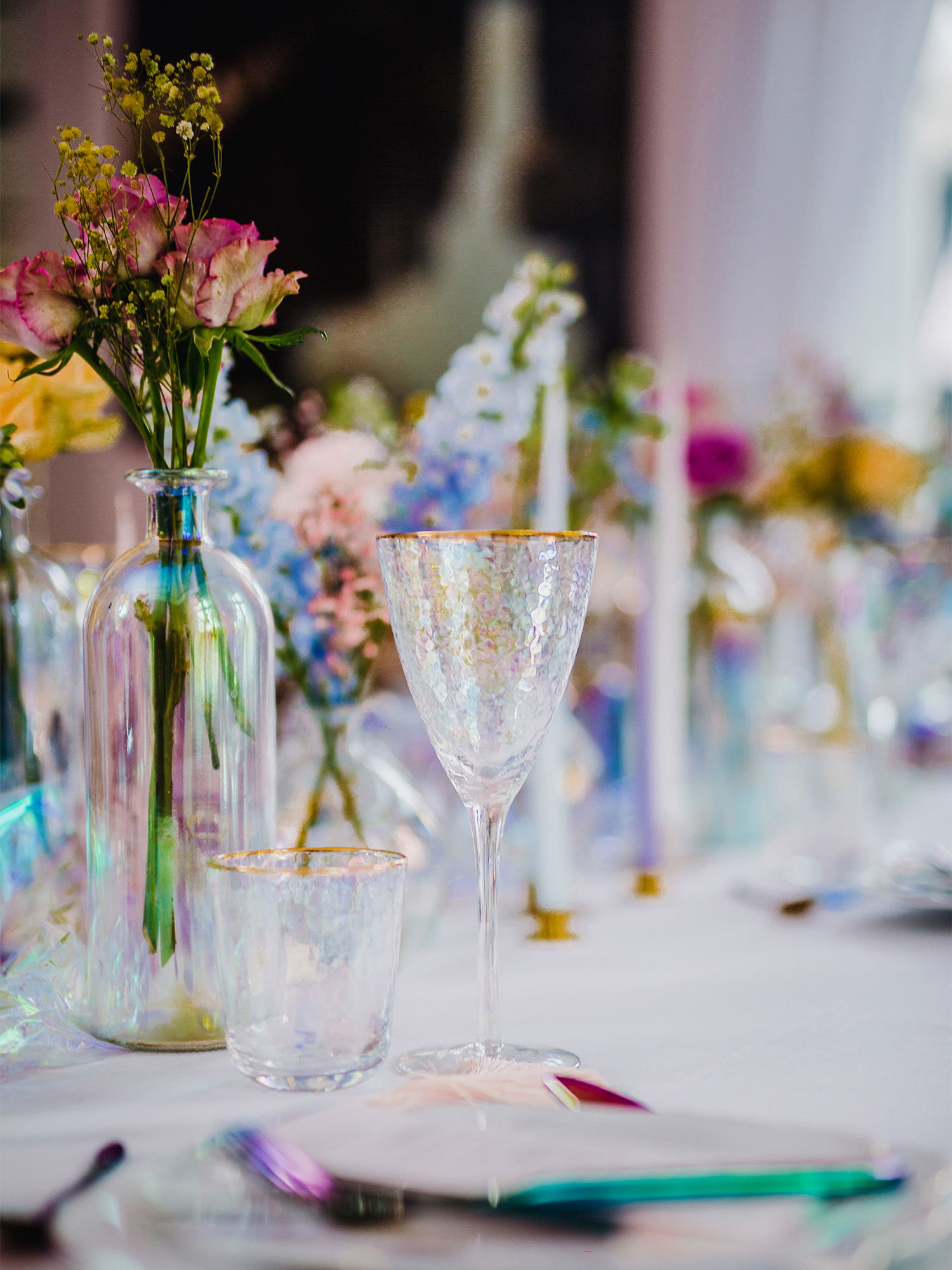Detailaufnahme eines festlich gedeckten Tisches mit schimmernden, facettierten Gläsern und einer Glasvase, die rosafarbene Blumen in die Tischdekoration integriert. Das Goldrand-Dekor der Gläser verstärkt die luxuriöse Atmosphäre, während die Blumenarrangements den Raum sanft erhellen.