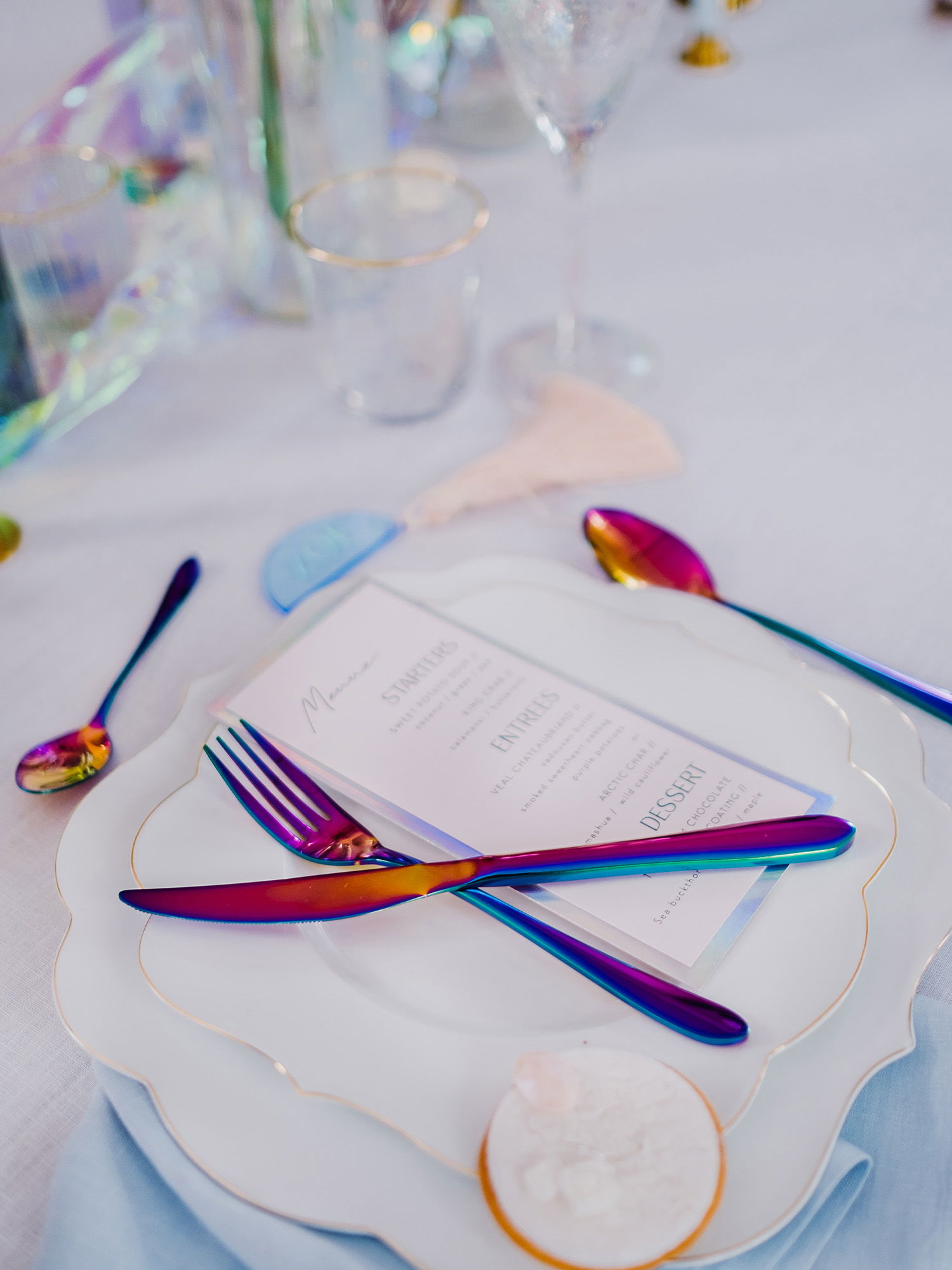 Detailansicht eines stilvoll gedeckten Tellers mit einem Menü. Das metallisch schimmernde Besteck und die dezente Goldverzierung des Tellers sorgen für ein luxuriöses Ambiente. Die sanften Farben der Tischdekoration ergänzen das edle Gesamtbild.