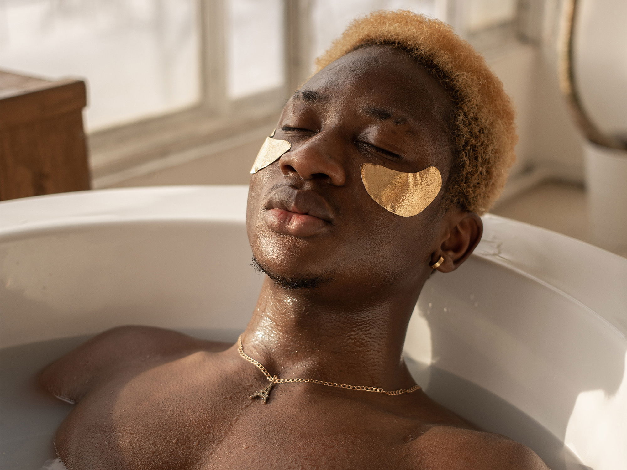 Ein Mann mit blonden Haaren und goldenen Augenmasken genießt ein entspannendes Bad. Das Licht, das durch das Fenster einfällt, erzeugt eine warme Atmosphäre. Seine nasse Haut glänzt leicht im Licht.