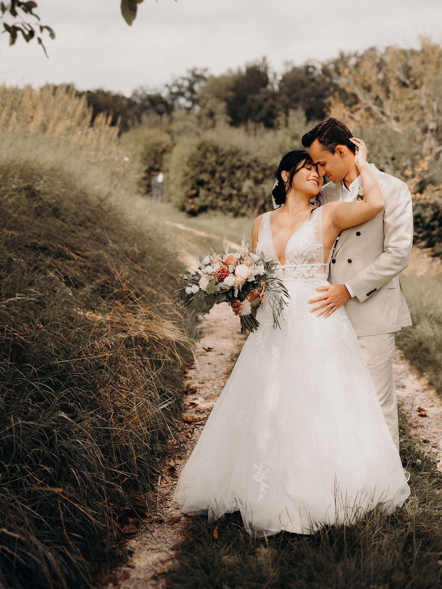 Eine Braut in einem weißen, fließenden Kleid steht auf einem Pfad neben ihrem Bräutigam im beigen Anzug. Sie lehnen sich aneinander, während sie lächelnd ihren Blumenstrauß hält.