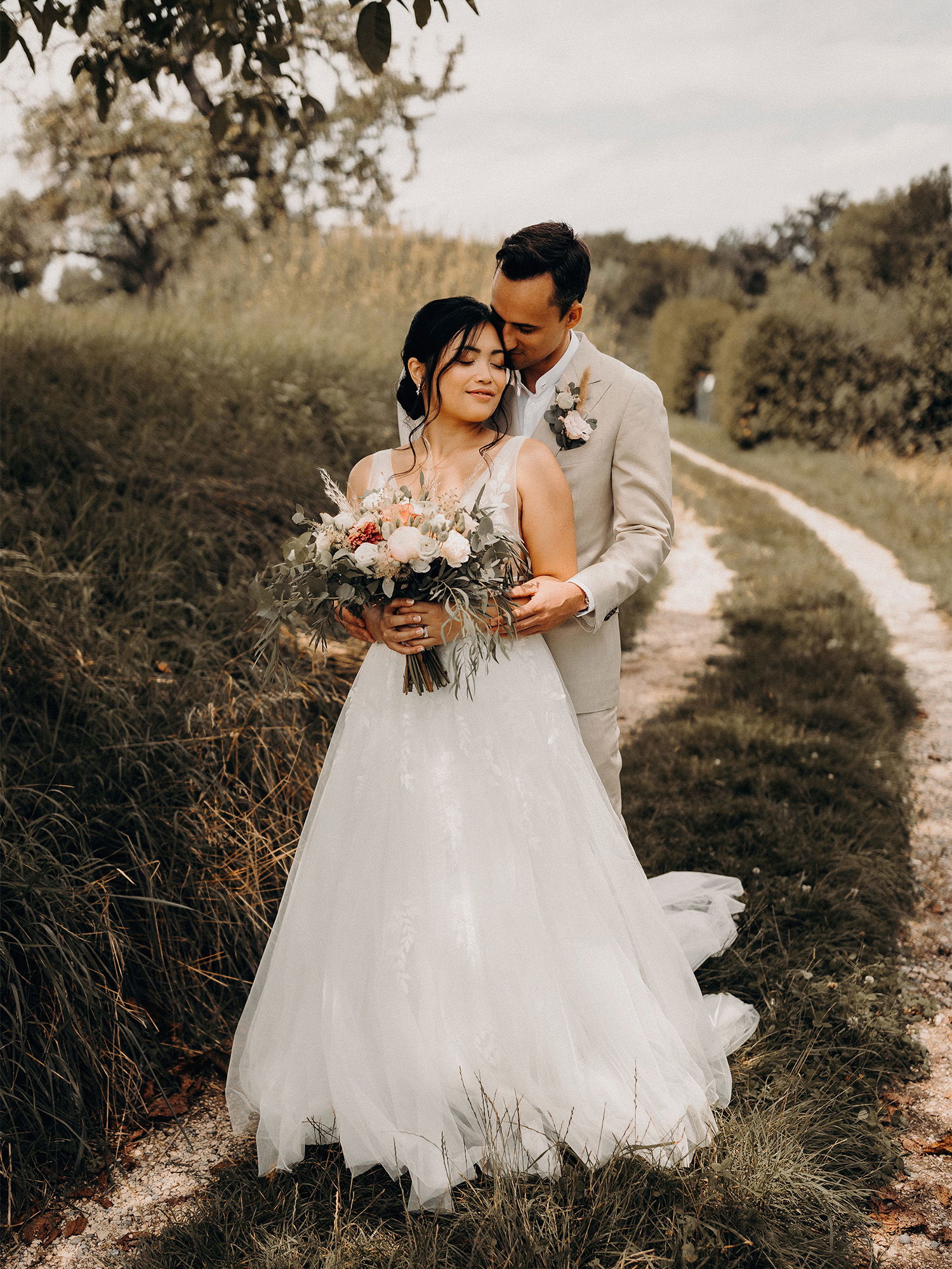 Der Bräutigam umarmt die Braut von hinten und küsst zärtlich ihren Kopf. Die Braut hält ihren Blumenstrauß vor sich, während sie entspannt die Augen schließt. Die Szene ist von Natur umgeben, mit Gras und Bäumen am Wegesrand.