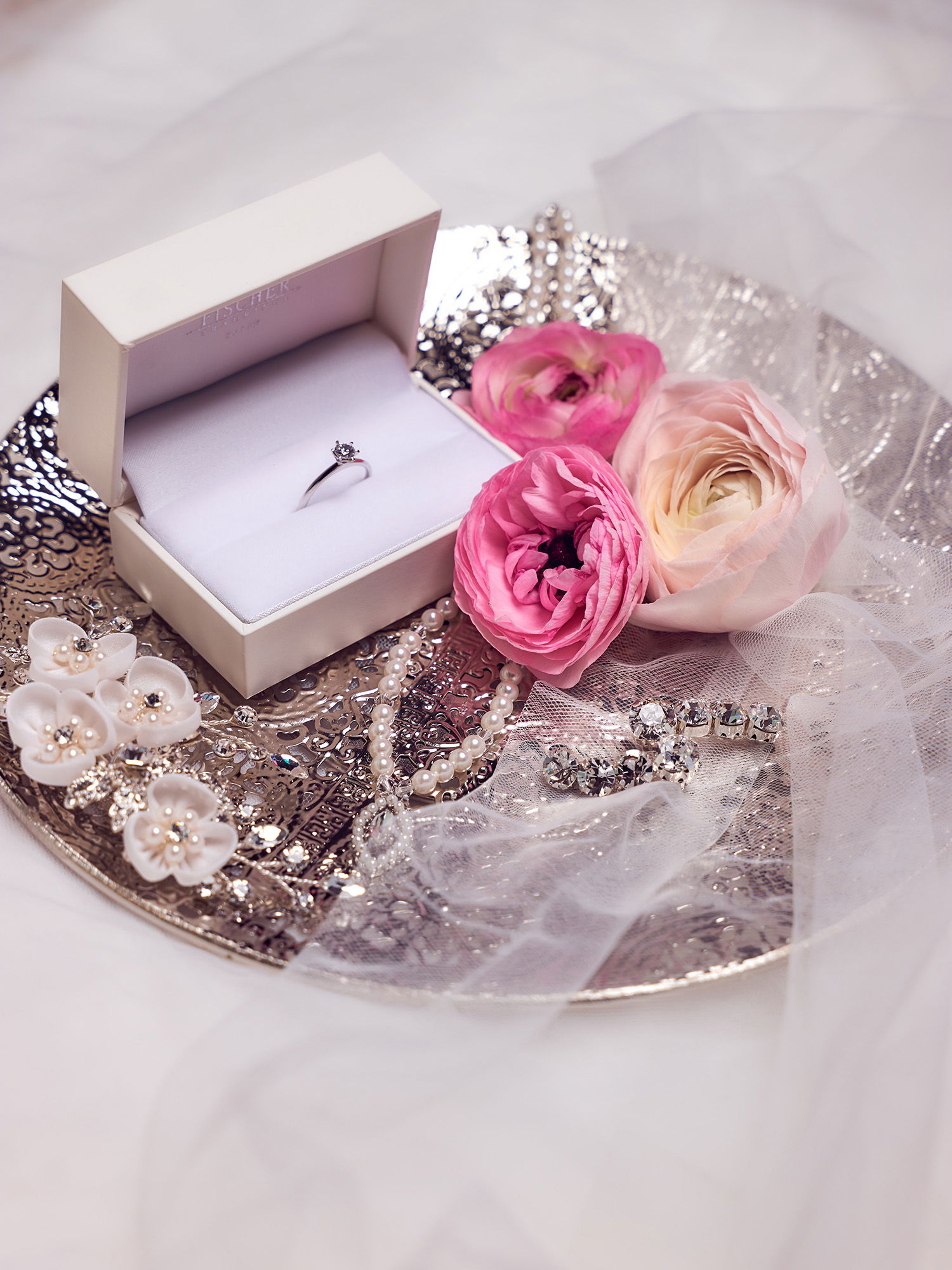 Ein Verlobungsring in einer offenen, weißen Box ruht auf einem kunstvoll verzierten Tablett. Um den Ring herum sind zarte, rosa Ranunkeln, Perlenschmuck und funkelnde Haarnadeln arrangiert. Der Schleier auf dem Tablett verstärkt den romantischen, festlichen Look.