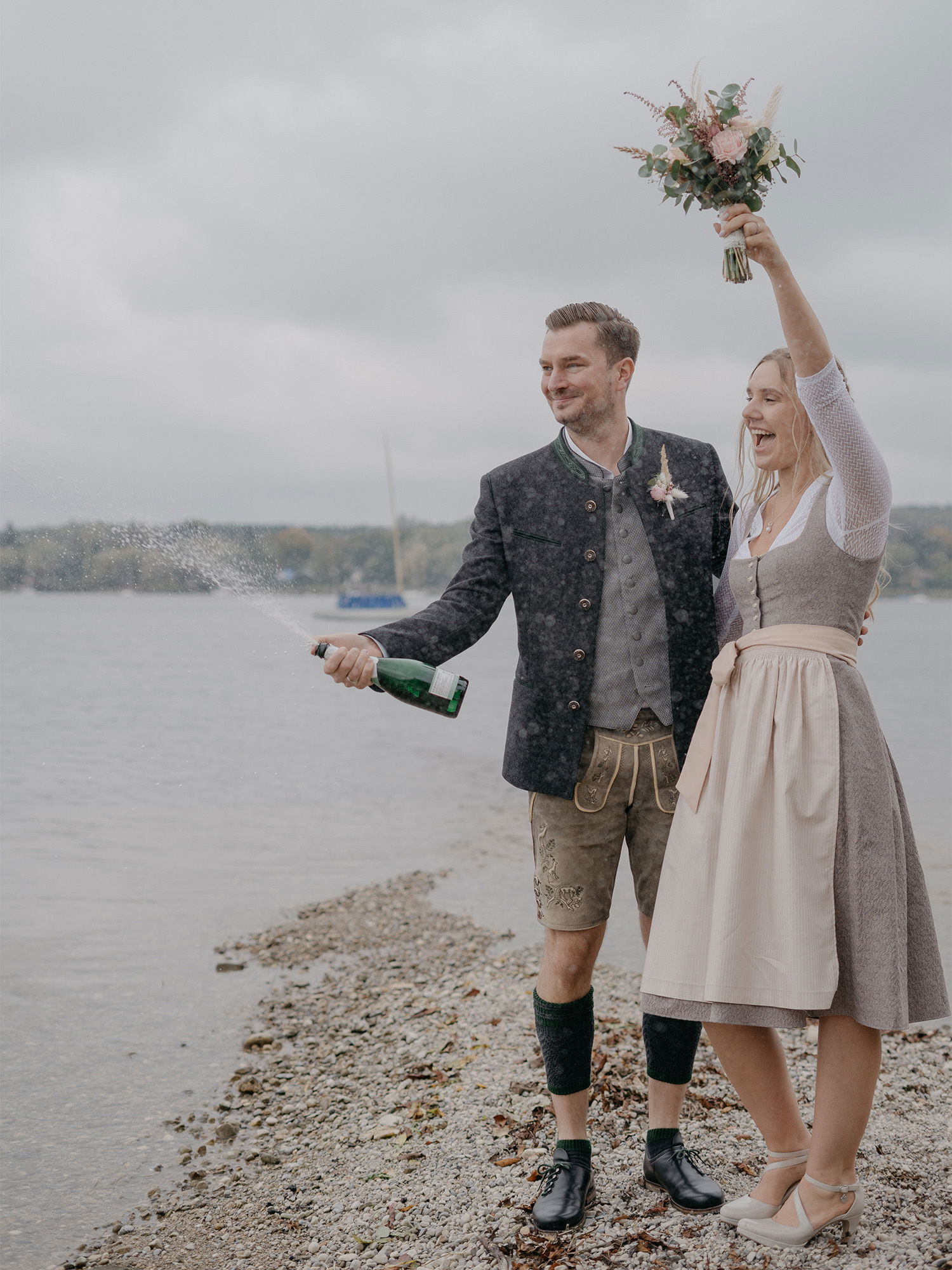 Ein traditionell bayerisch gekleidetes Paar feiert am Ufer eines Sees. Der Bräutigam, in Lederhosen und Janker, sprüht Sekt, während die Braut mit erhobenem Arm und Blumenstrauß jubelt.