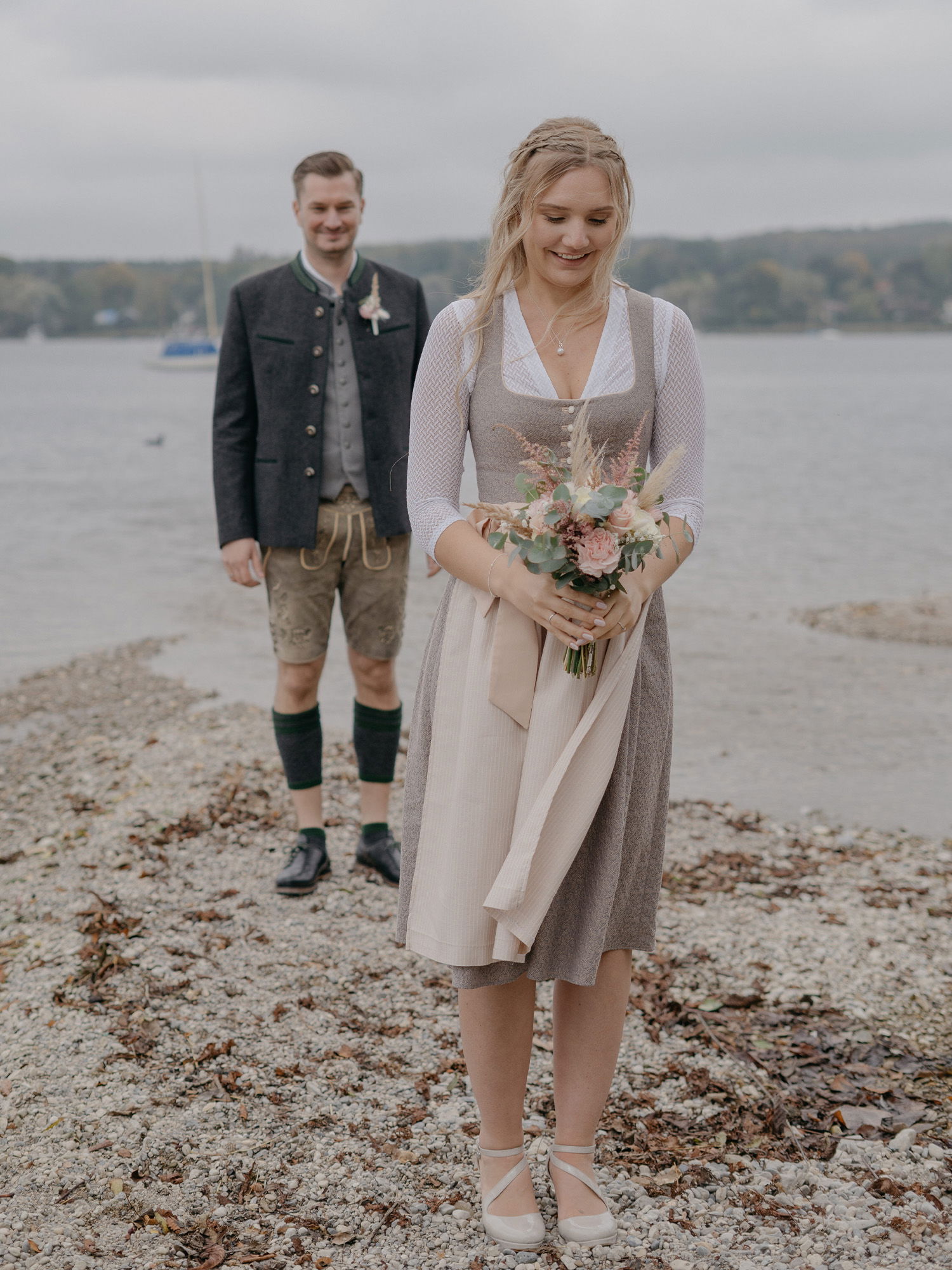 Die Braut, in einem klassischen Dirndl, steht am See und blickt sanft auf ihren Blumenstrauß, während der Bräutigam lächelnd im Hintergrund steht.