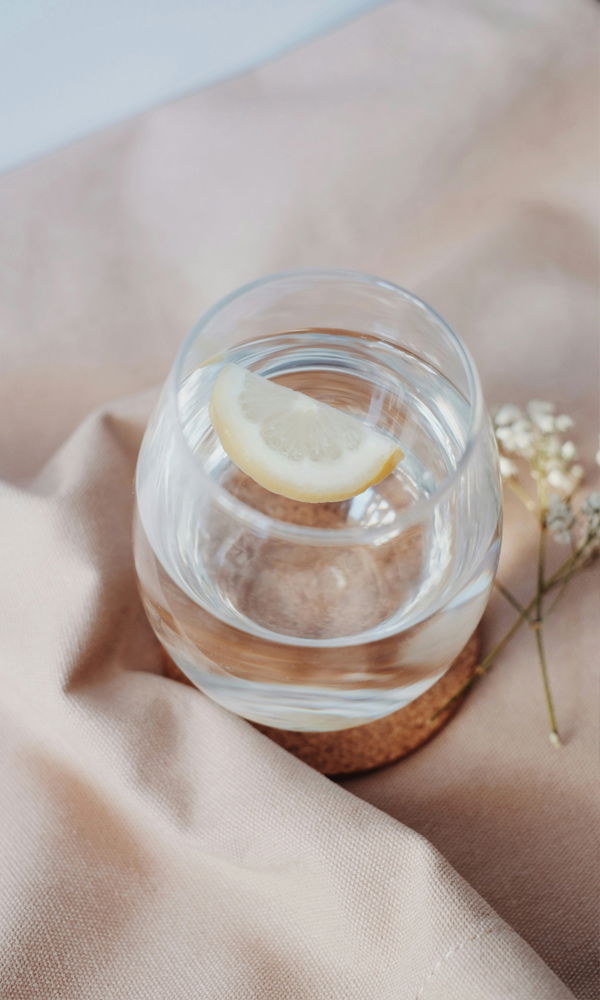 Frisches Wasser mit einer Zitronenscheibe in einem transparenten Glas, dekoriert mit sanftem Stoff und kleinen Blumenakzenten im Hintergrund.