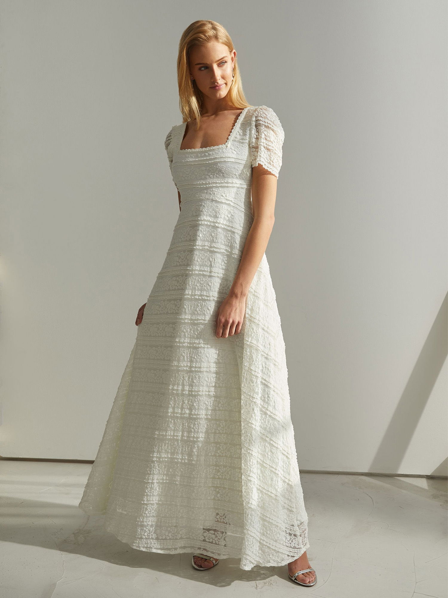 Eine Braut in einem eleganten, weißen Kleid mit strukturiertem Stoff und kurzen Ärmeln posiert in einem schlichten Raum. Der klare Schnitt des Kleides und der weiche Lichteinfall betonen den modernen Look.