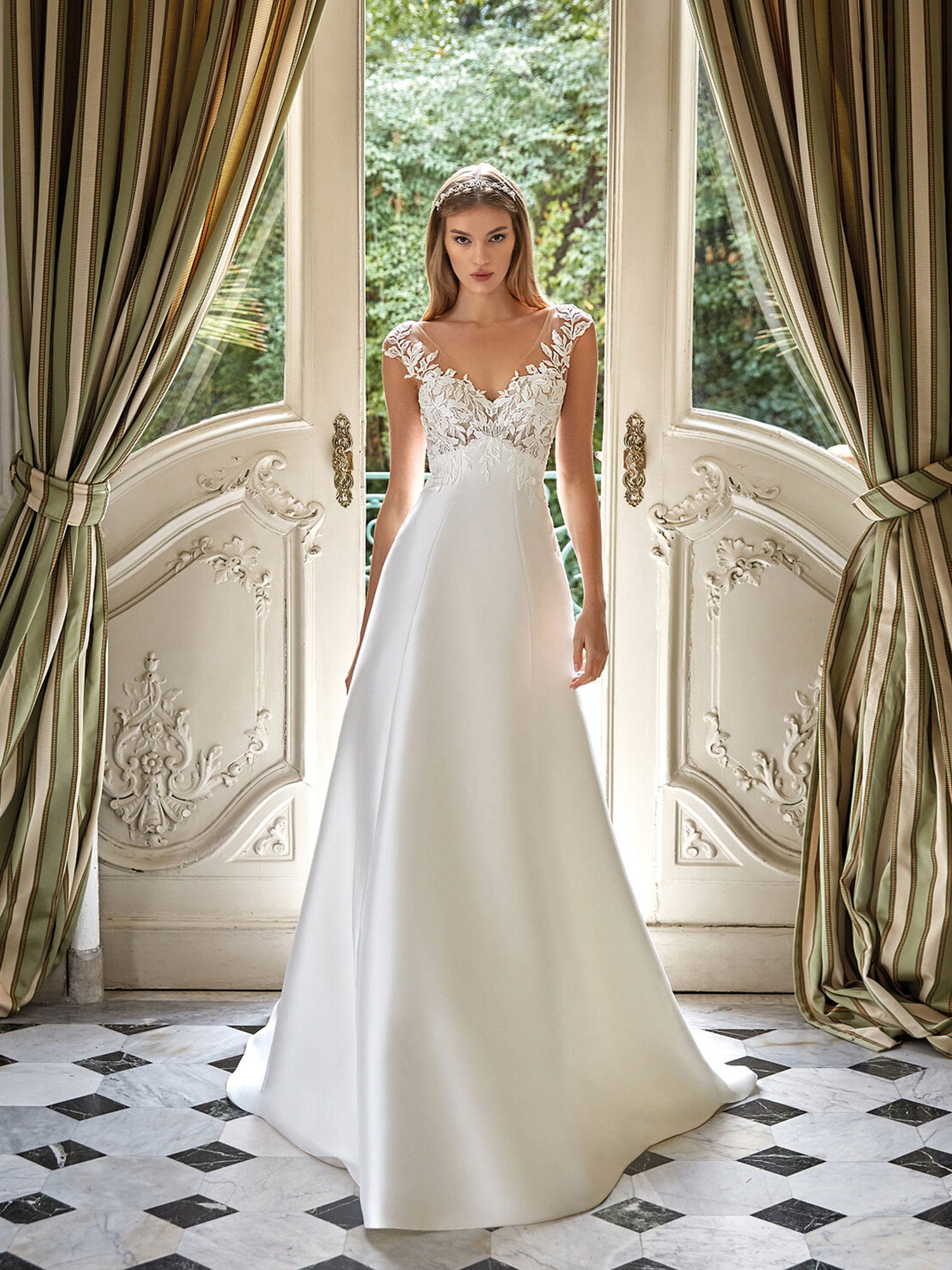 Eine Frau steht in einem opulenten Raum und trägt ein elegantes, bodenlanges Brautkleid mit einem Spitzenoberteil und einem schlichten Rock. Die geöffneten Türen hinter ihr führen auf einen grünen Garten, was der Szene eine verträumte Atmosphäre verleiht.