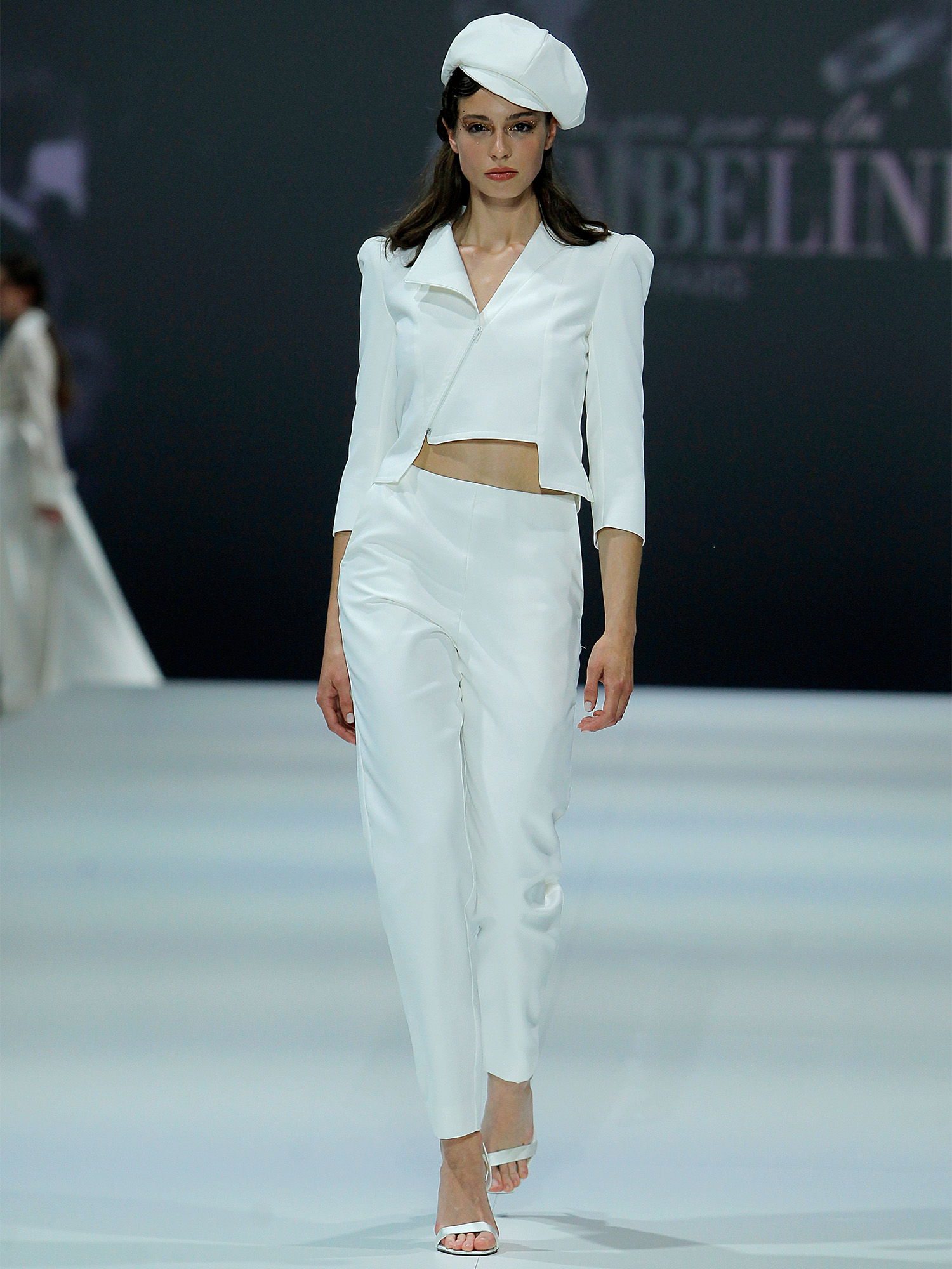 Auf einem Laufsteg trägt ein Model einen weißen Zweiteiler mit einem kurzen Blazer und einer schmal geschnittenen Hose. Sie kombiniert das Outfit mit einer stilvollen Mütze, was dem Look eine moderne und schicke Note verleiht.
