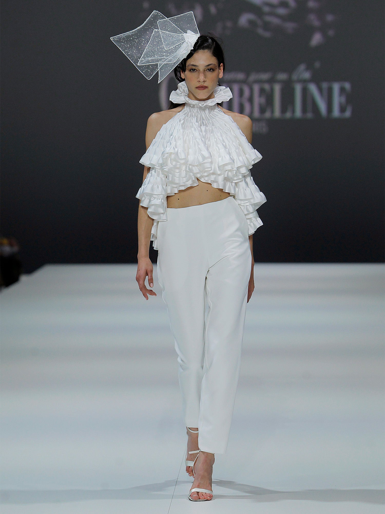 Eine Frau auf dem Laufsteg trägt ein Brautoutfit, bestehend aus einem rüschenverzierten Oberteil und einer schlichten, eleganten Hose. Auf ihrem Kopf trägt sie einen auffälligen Kopfschmuck, der das modische und kreative Design des Outfits unterstreicht.