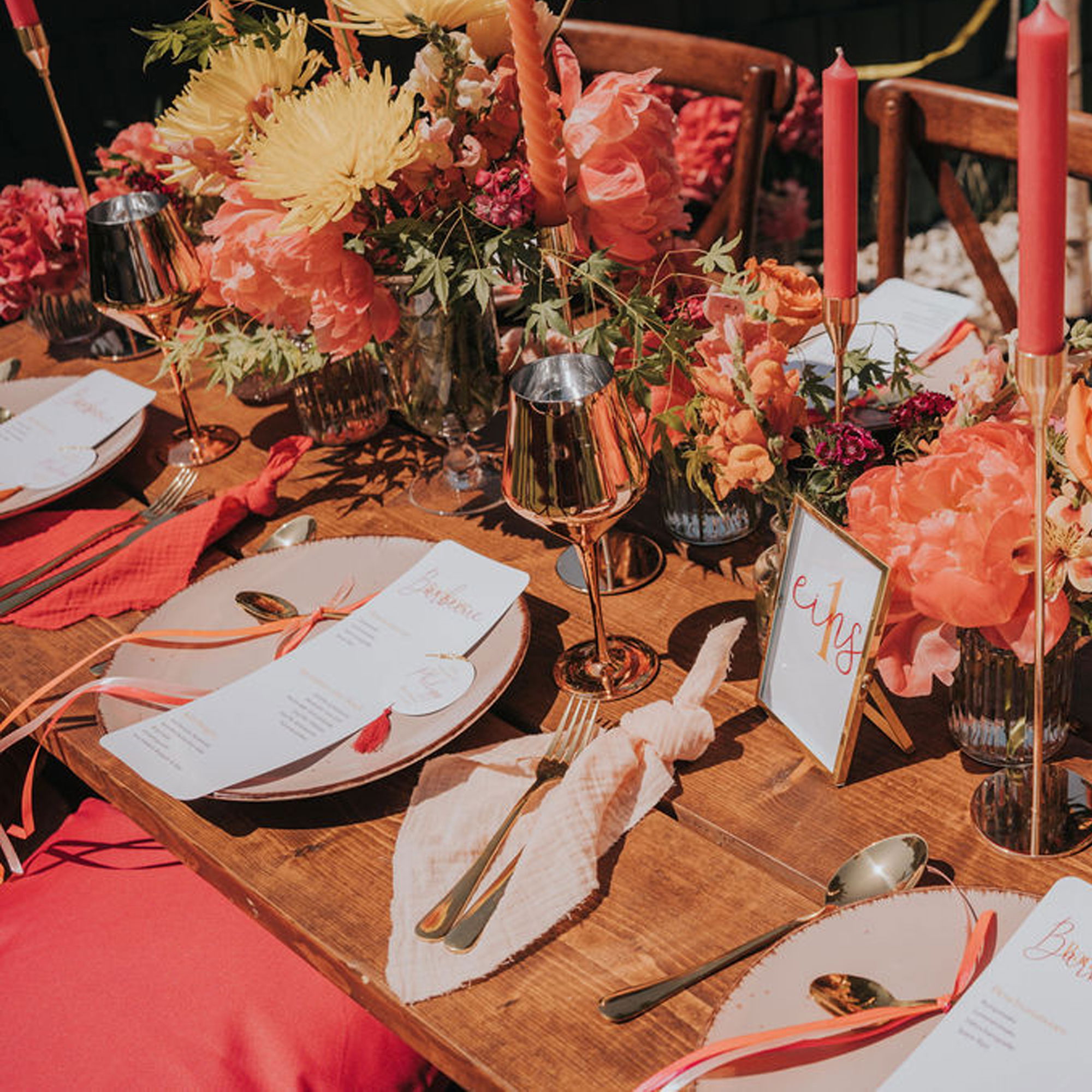 Festlich gedeckter Holztisch mit goldenen Bestecken, orangefarbenen Kerzen, farbenfroher Blumendekoration und individuell gestalteten Platzkarten.