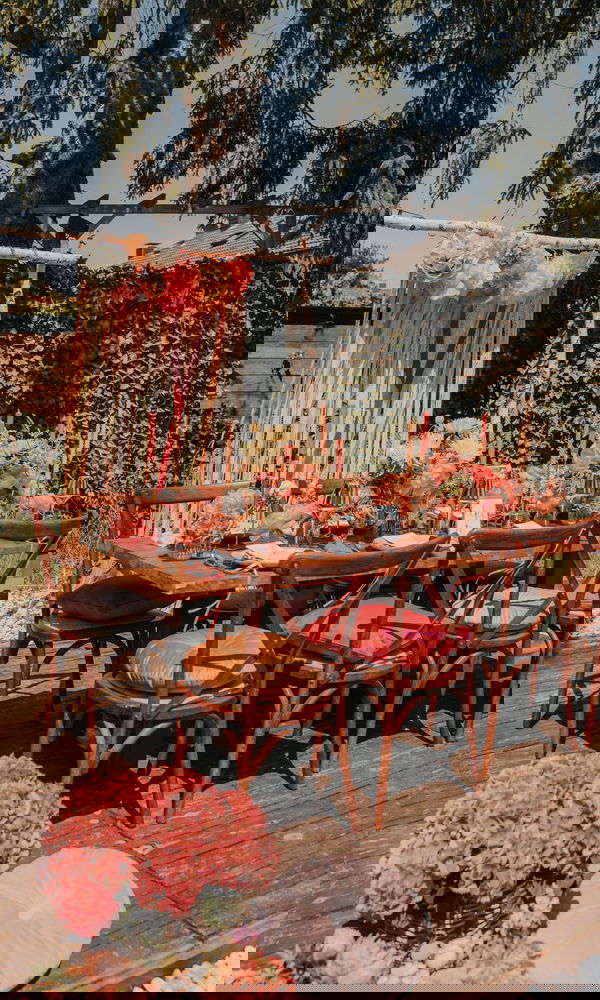 Festlich gedeckte Tafel für eine Gartenhochzeit mit Holzstühlen, üppigen Blumengestecken in warmen Farben und Akzenten wie Kerzen und Kissen.