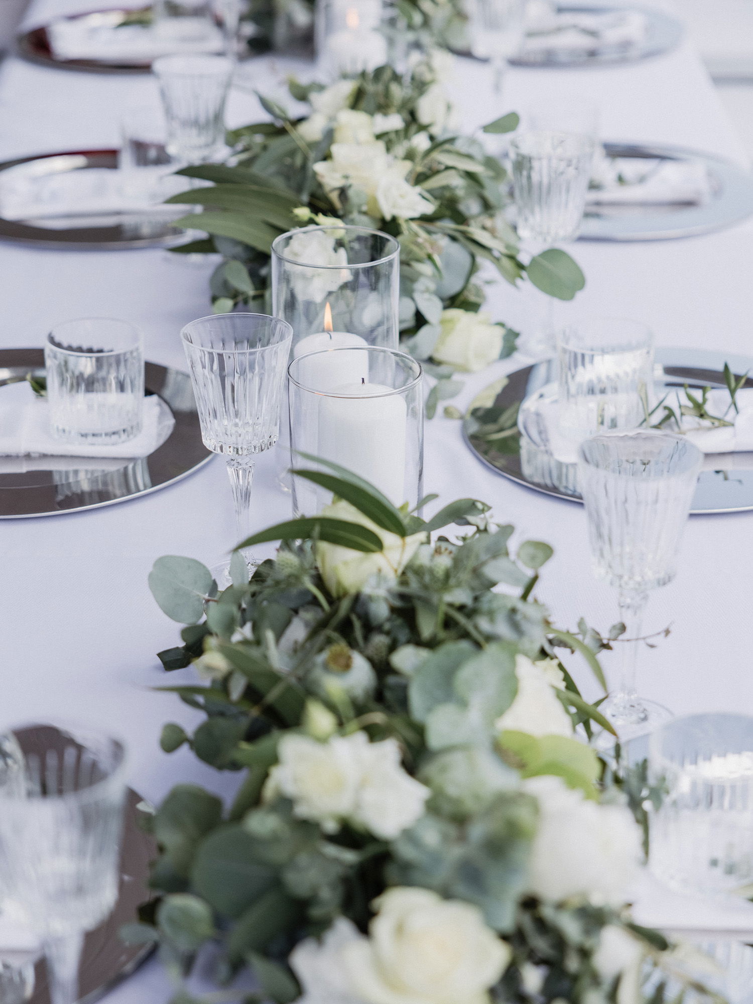 Detailaufnahme einer Hochzeitstafel, auf der Kerzen in Gläsern und üppige Blumenarrangements mit Eukalyptusblättern die Dekoration abrunden. Im zweiten Bild hält die Braut einen Brautstrauß aus Eukalyptusblättern und zarten weißen Blumen, während sie über ein Grasfeld geht.